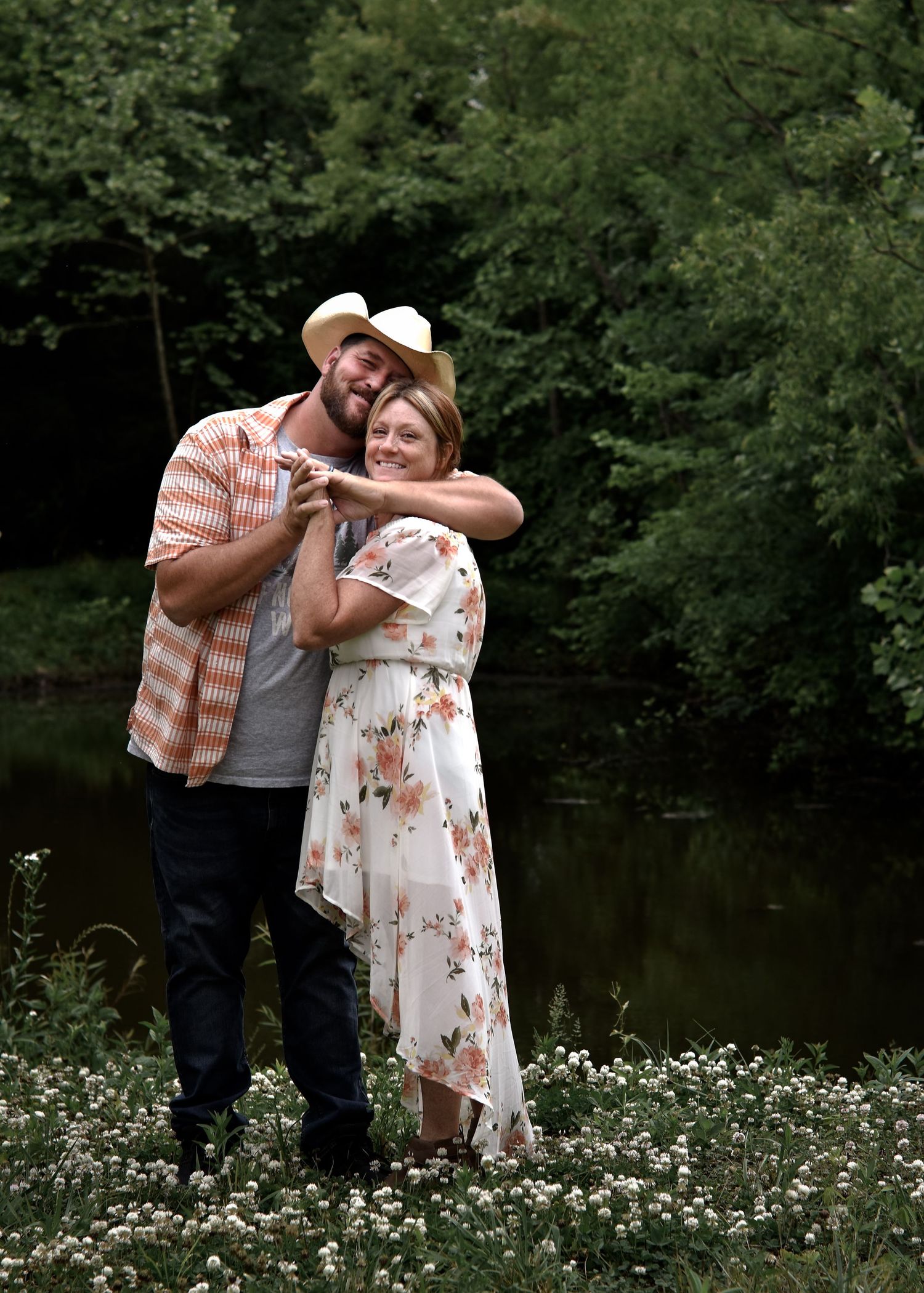 Casual wedding couple with groom in cowboy hat and bride in floral dress, holding hands by pond with lush greenery, in north Alabama