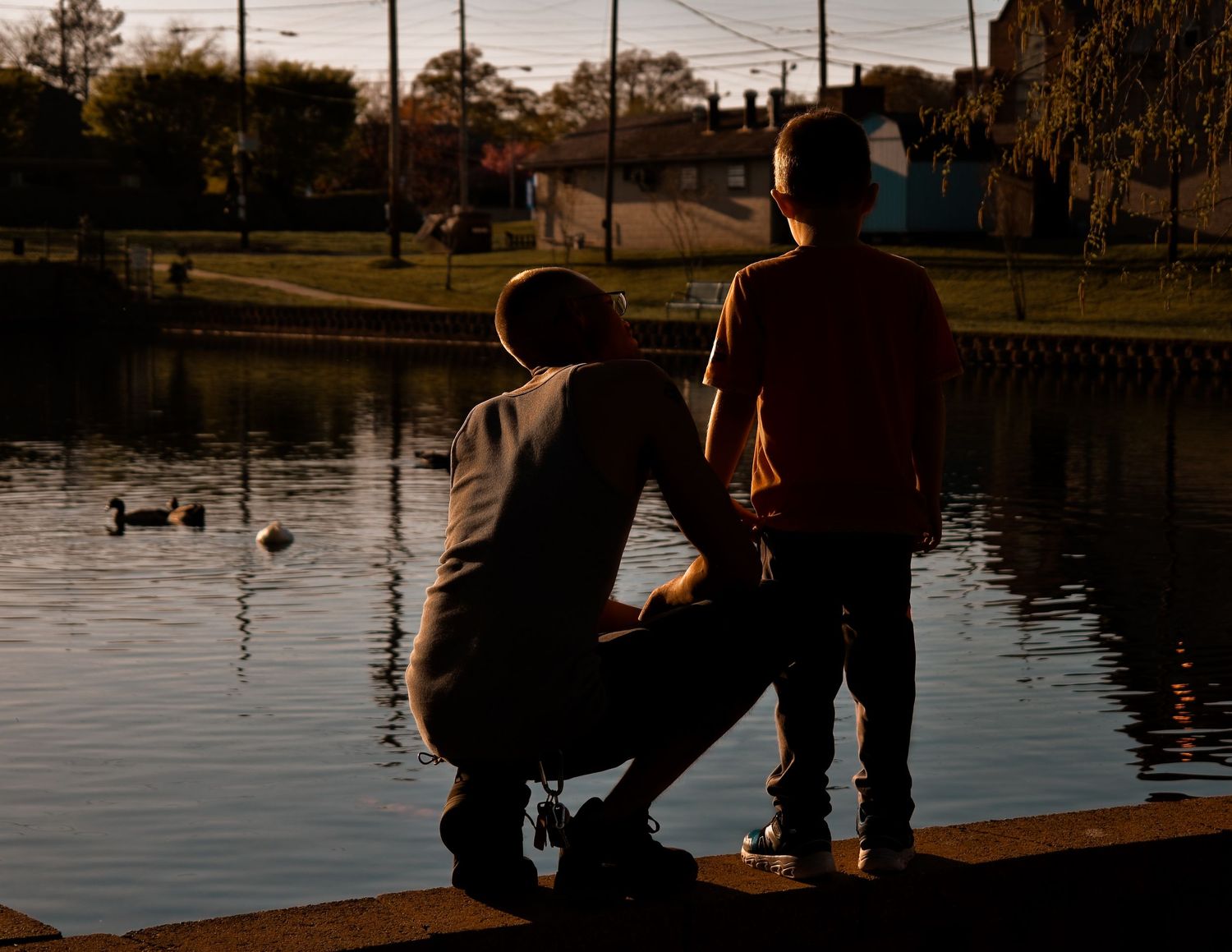 Emotional and candid silhouette of father kneeling beside son at the edge of a duck pond at sunset captured by Catch Your Fire Photography.