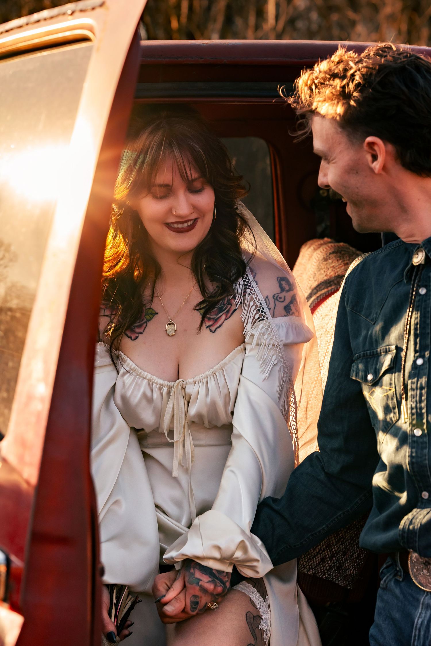 Bride sitting in driver’s seat of red truck, blushing and smiling, groom looking at her, sun glowing over door. Huntsville, Alabama.