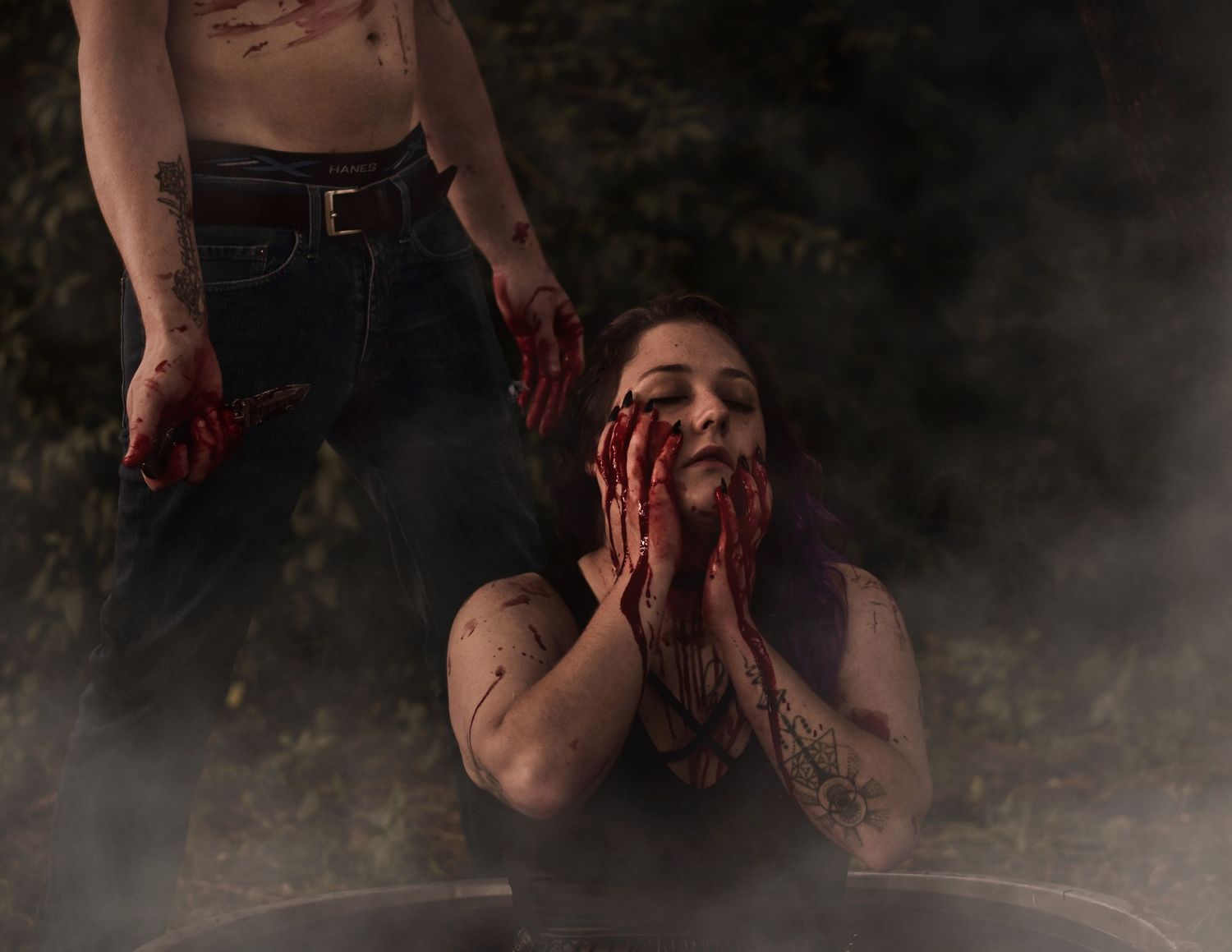 creative editorial horror image of girl bathing in fake blood as boyfriend stands behind with prop knife ready to end it all
