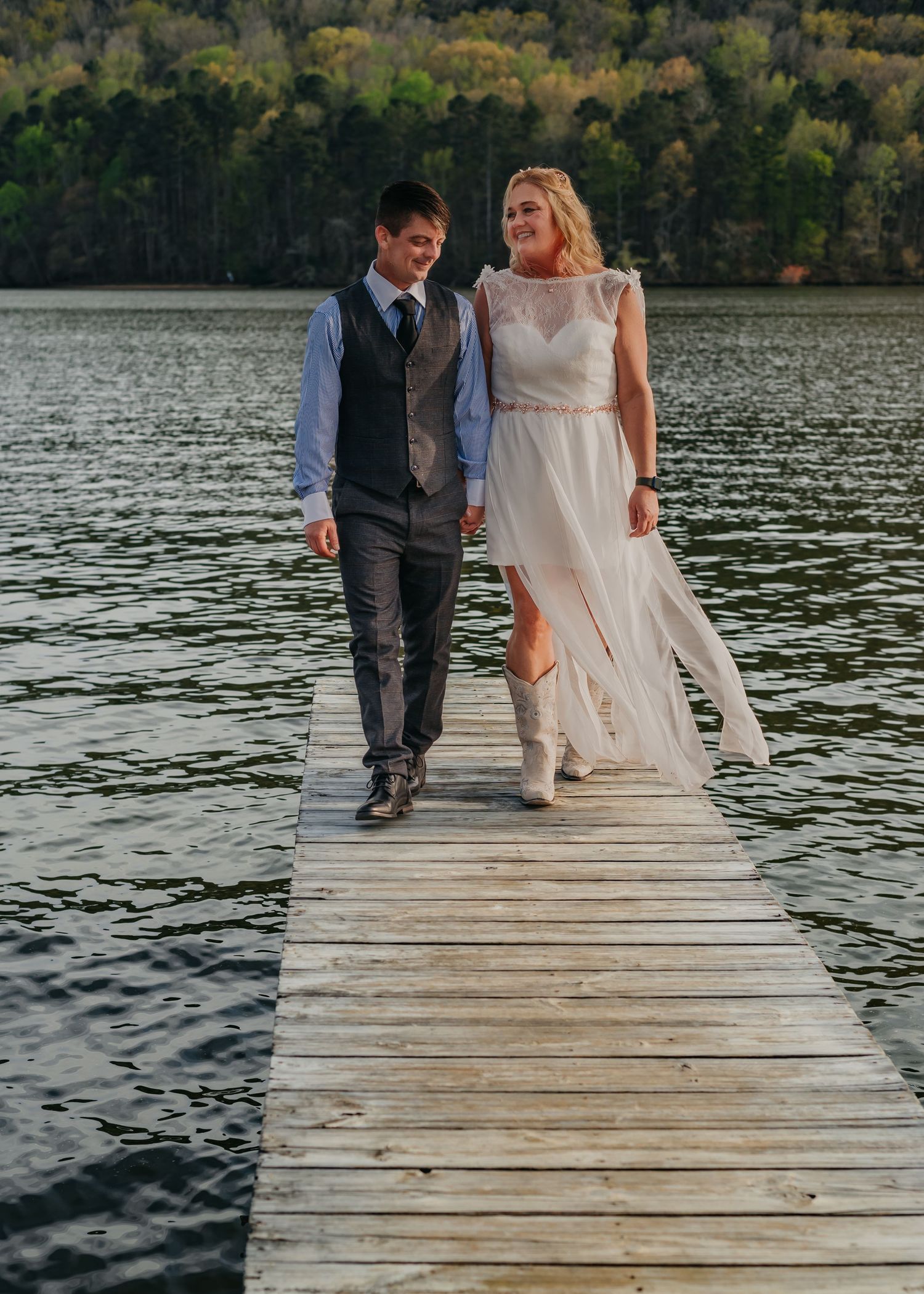 Bride and groom holding hands walking toward camera on wooden dock at spring wedding by river, water and trees in background
