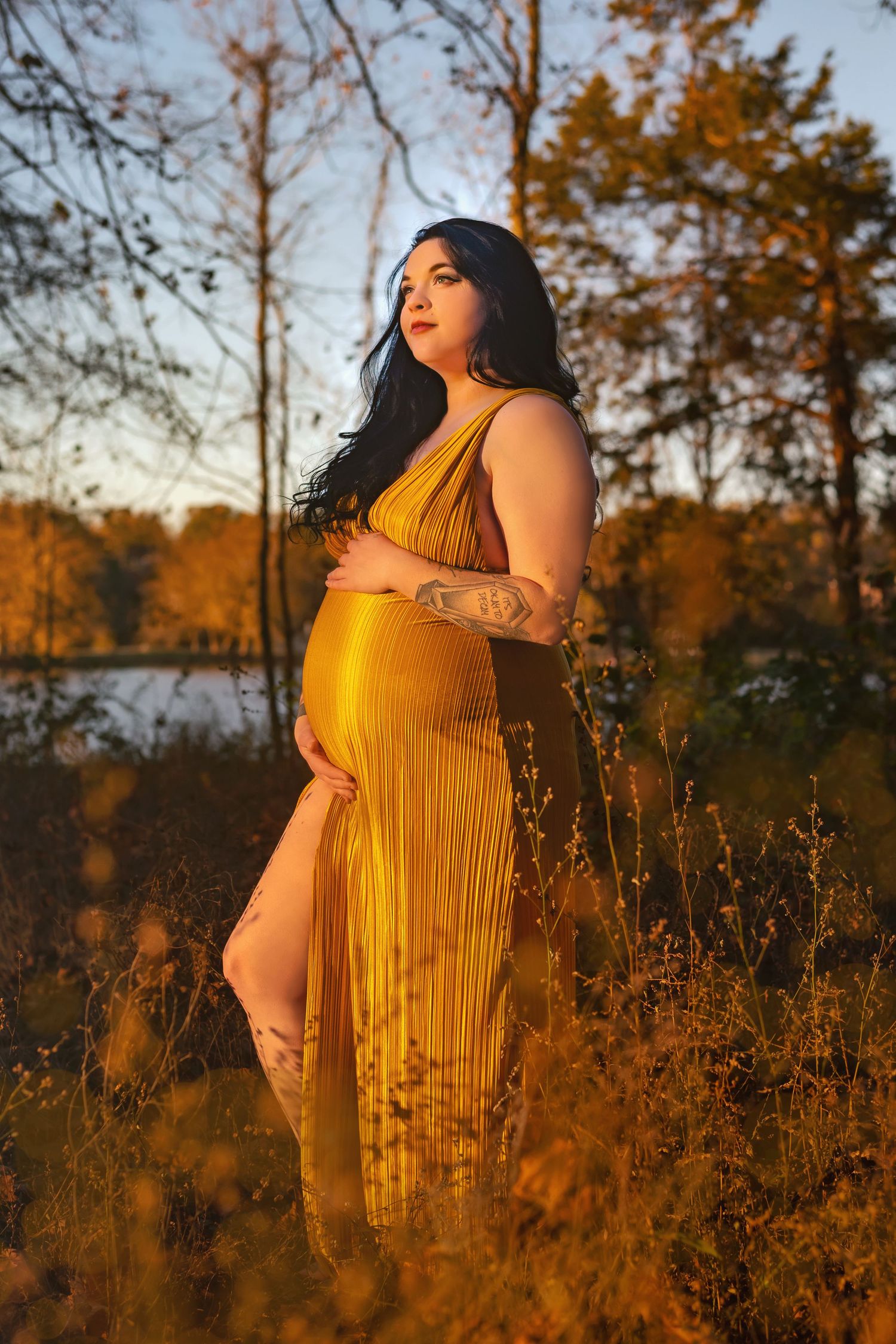 Warm maternity session with woman in yellow dress, sunlit glow, lake and trees blurred behind, captured by Catch Your Fire Photography.