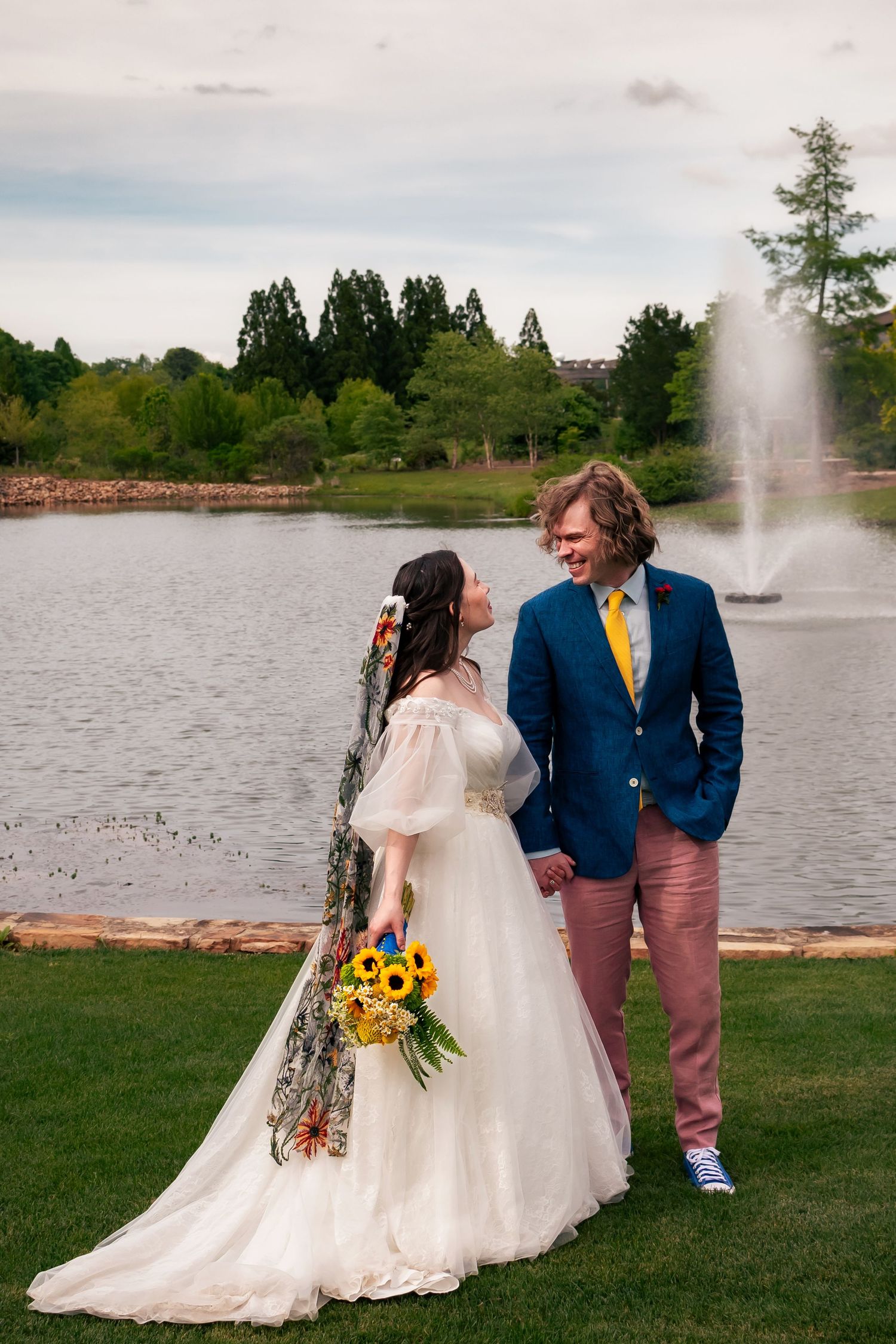 Cinematic, joyful wedding moment of bride and groom walking hand in hand by pond with fountain at Huntsville Botanical Garden wedding.
