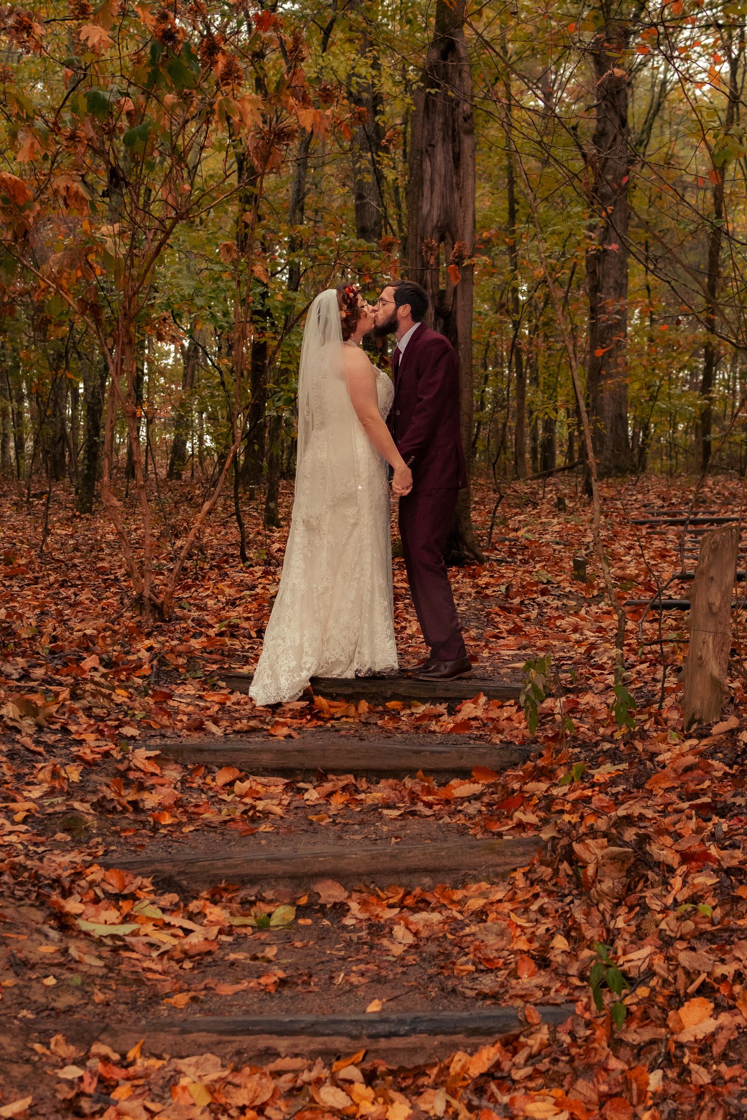 Cinematic wedding shot of bride and groom kissing on leaf-covered forest steps at Madison County Nature Trail. Like a dreamy storybook.