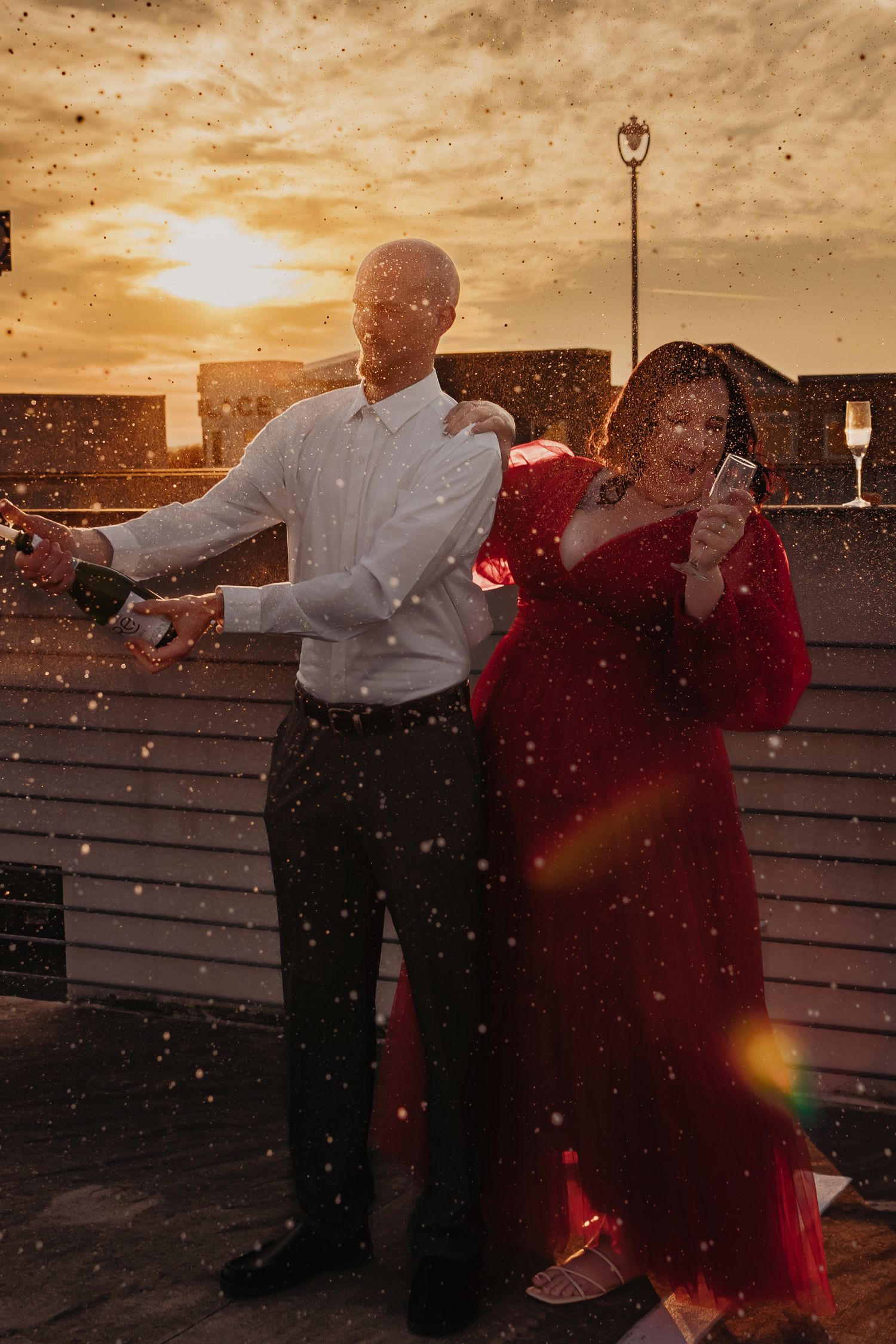 Bridge Street engagement rooftop at sunset, bride in red gown laughing behind groom as champagne pops and sprays.