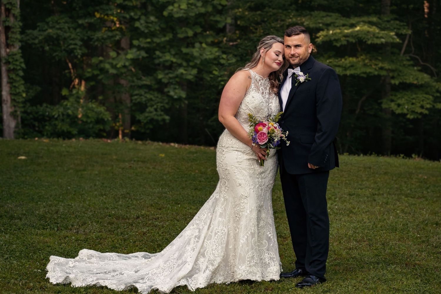 Bride and groom standing in front of green trees as bride leans her head on the groom with closed eyes and a content smile at Ivy Oaks venue