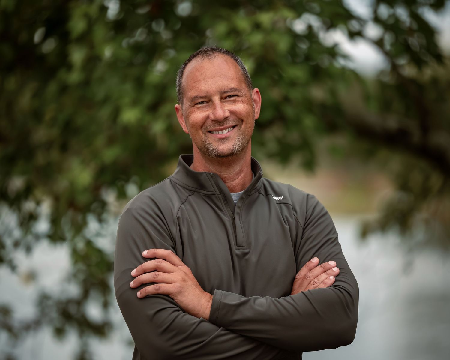 Casual headshot portrait of man with arms crossed, smiling at camera, tree leaves and lake softly blurred in background at Edgewater Park.