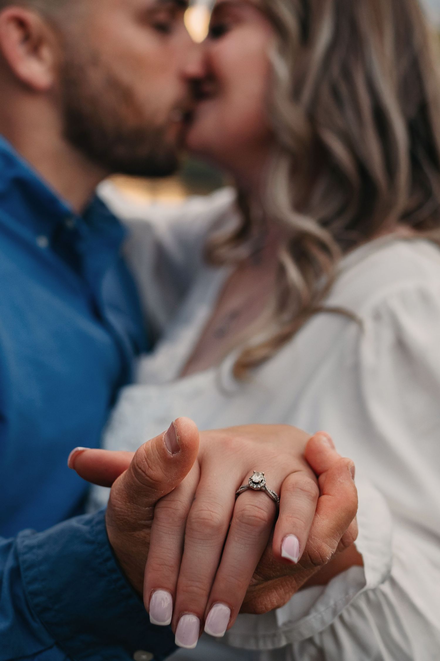 Engagement ring detail shot with couple kissing blurred in background, his hand holding hers, captured by Catch Your Fire Photography.