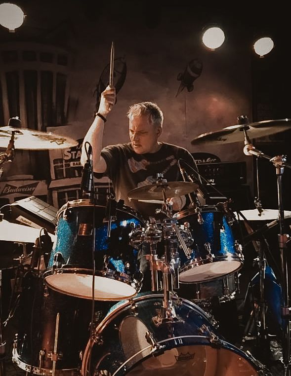 Live show shot of drummer in action with stick in air, lights glowing in back, playing blue glitter drum set at Huntsville, Alabama concert.