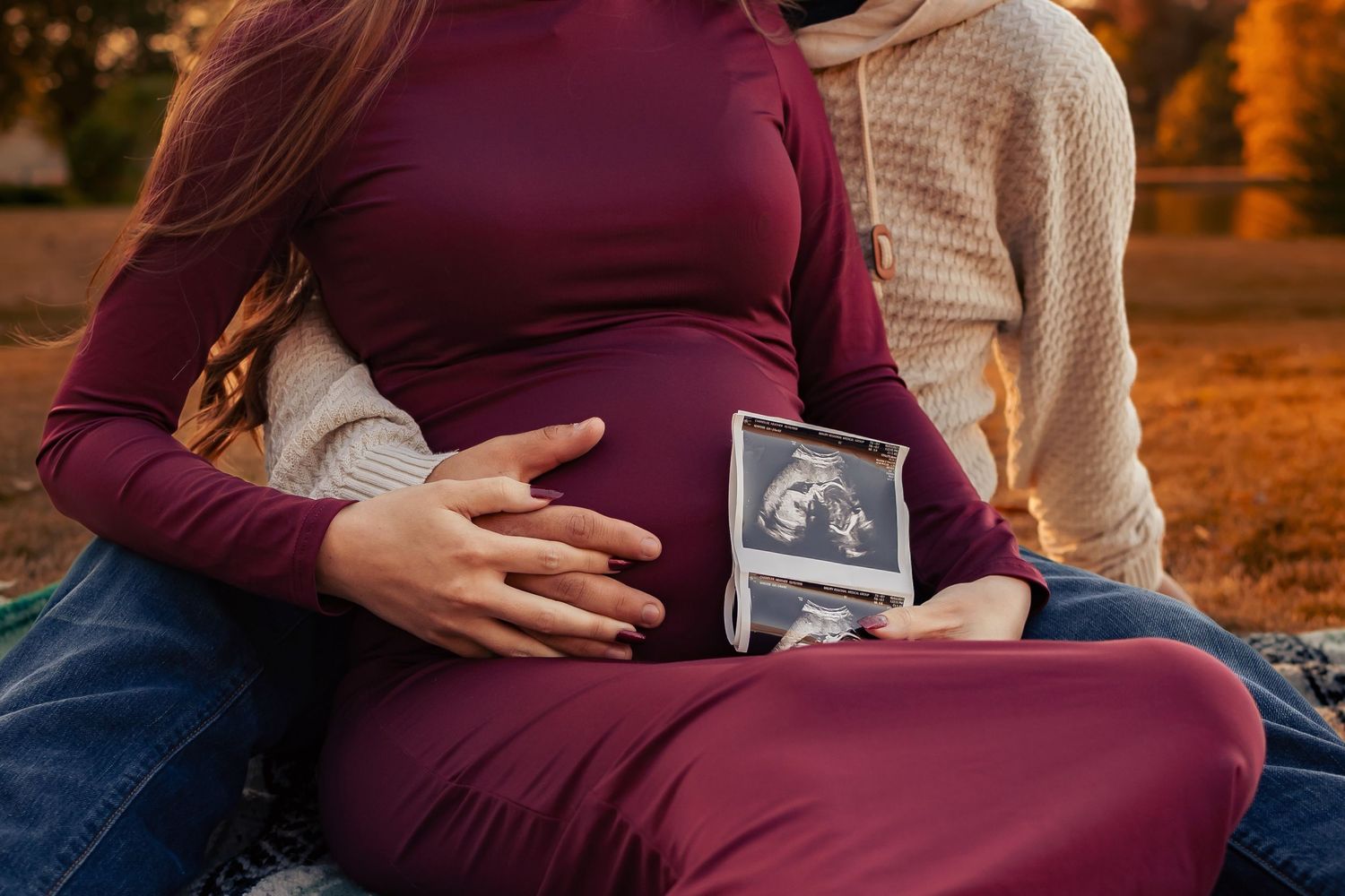 closeup maternity detail image of mom and dad siting on the ground holding belly and ultrasound photo by Lady Ann Lake in Huntsville, AL