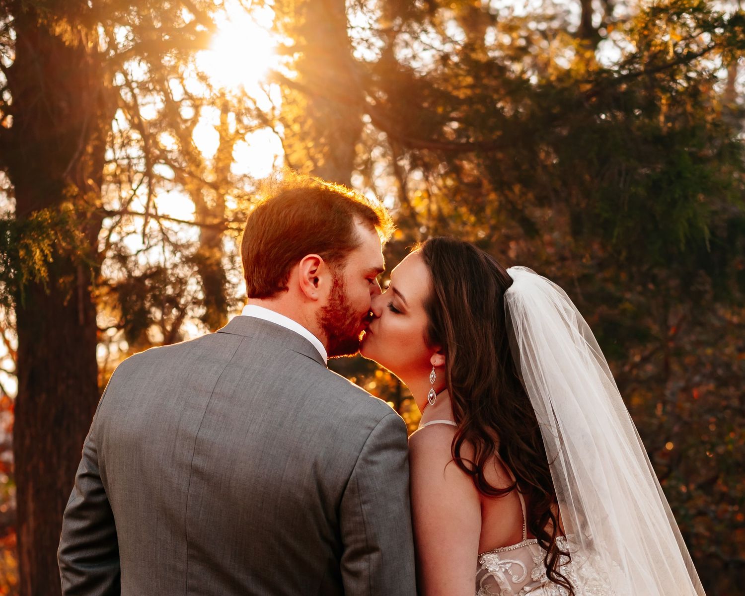 Couple sharing a kiss, backs to camera, with sun glowing through trees, photographed by Catch Your Fire Photography in Huntsville, AL.