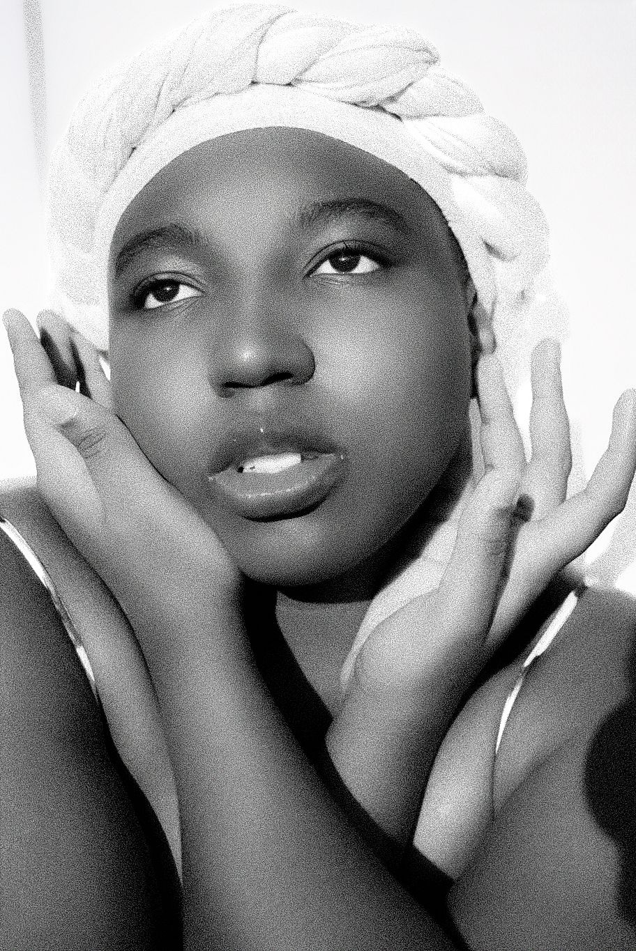 Black and white portrait of a young woman with a braided towel wrap on her head, hands framing her face.