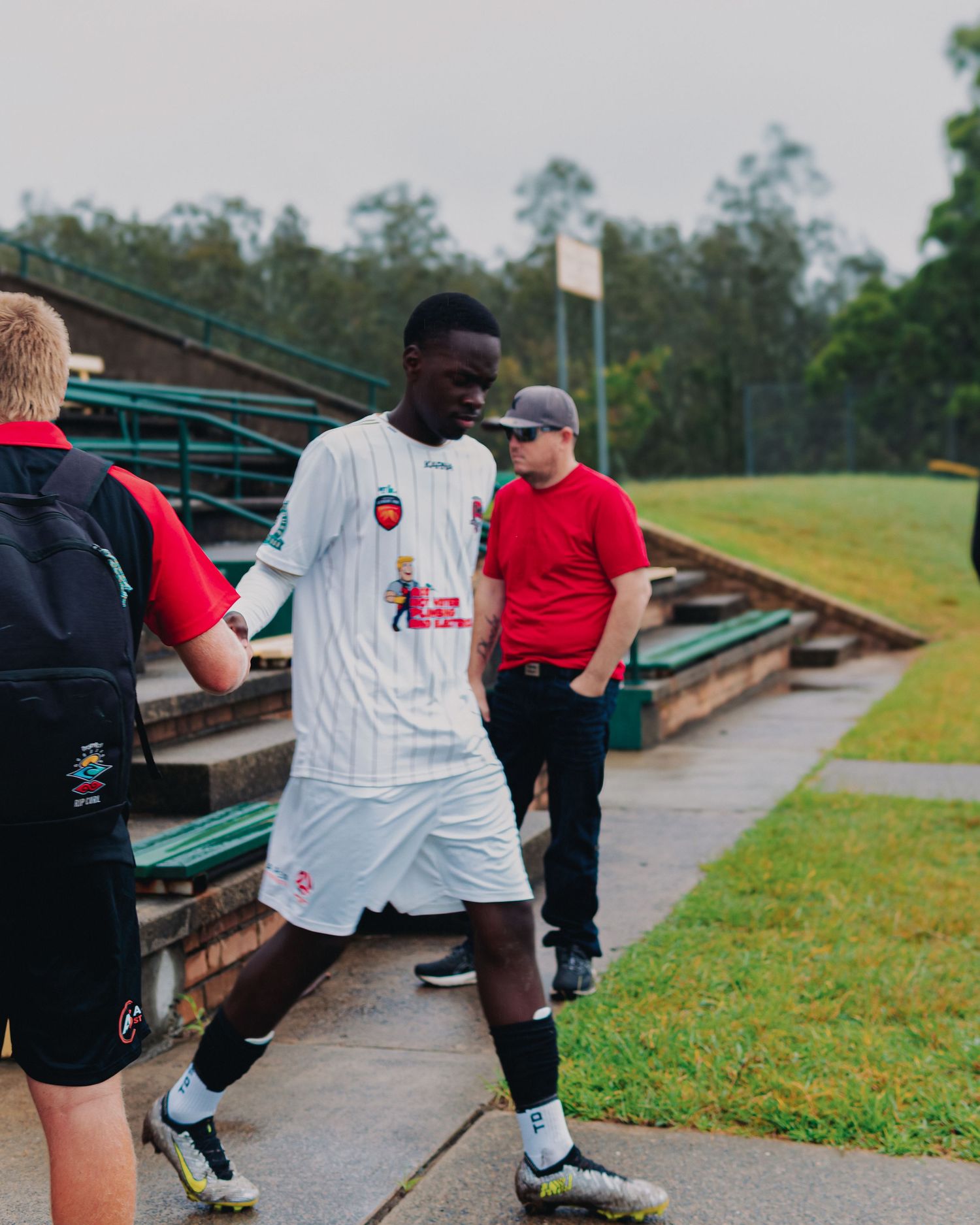 Soccer player in white uniform walks near stadium bleachers with teammates and staff on a grassy field.