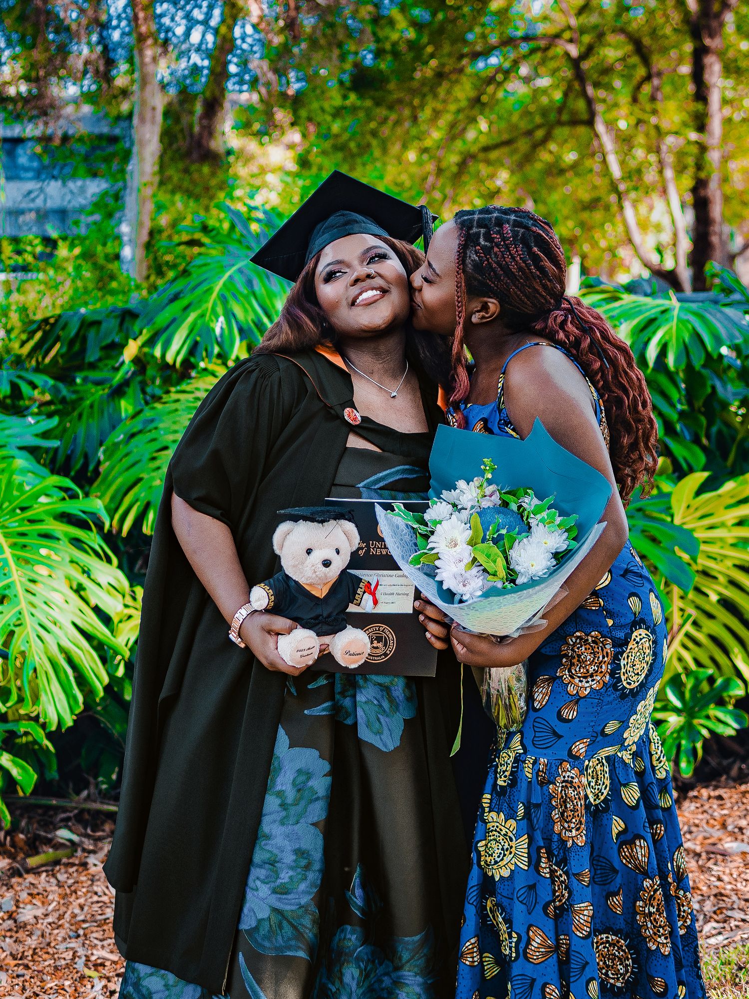 Graduate in cap and gown receives kiss while holding teddy bear and flowers.
