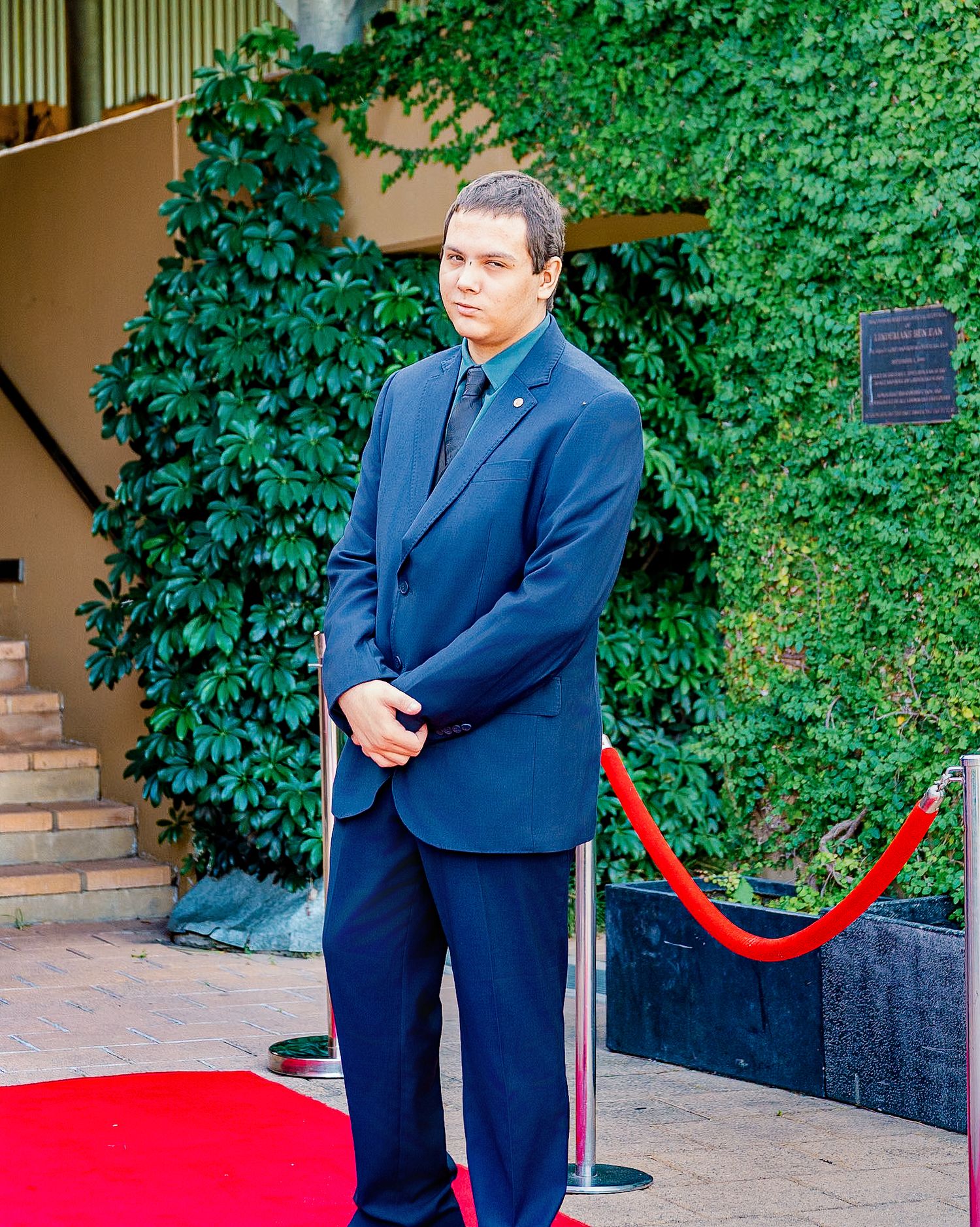 Man in blue suit standing on red carpet near velvet rope with lush green ivy wall backdrop.
