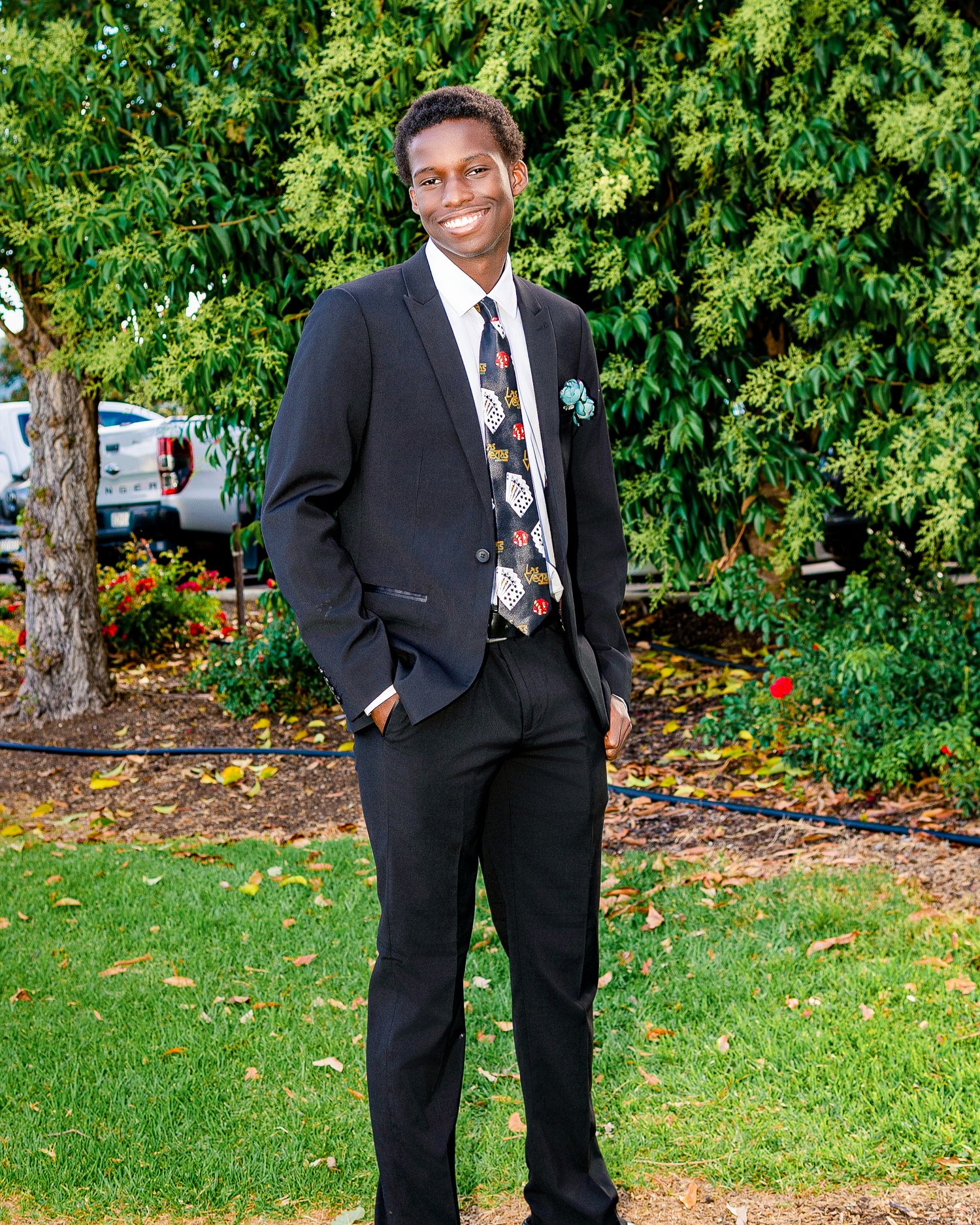 Smiling young man in black suit with floral tie and pocket square poses outdoors in garden.