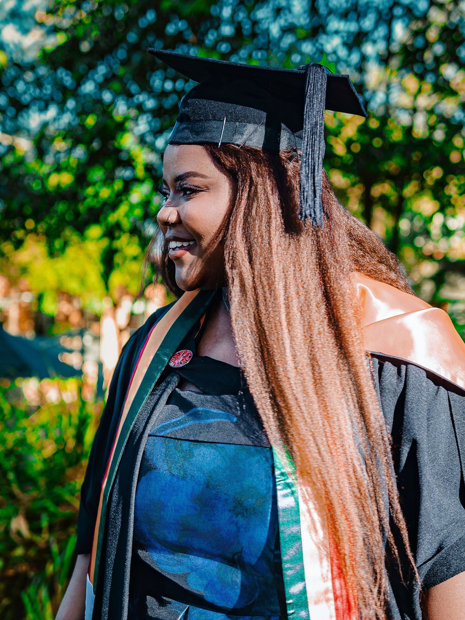 Smiling graduate in black cap and gown with colorful sash laughs joyfully in sunny outdoor setting.
