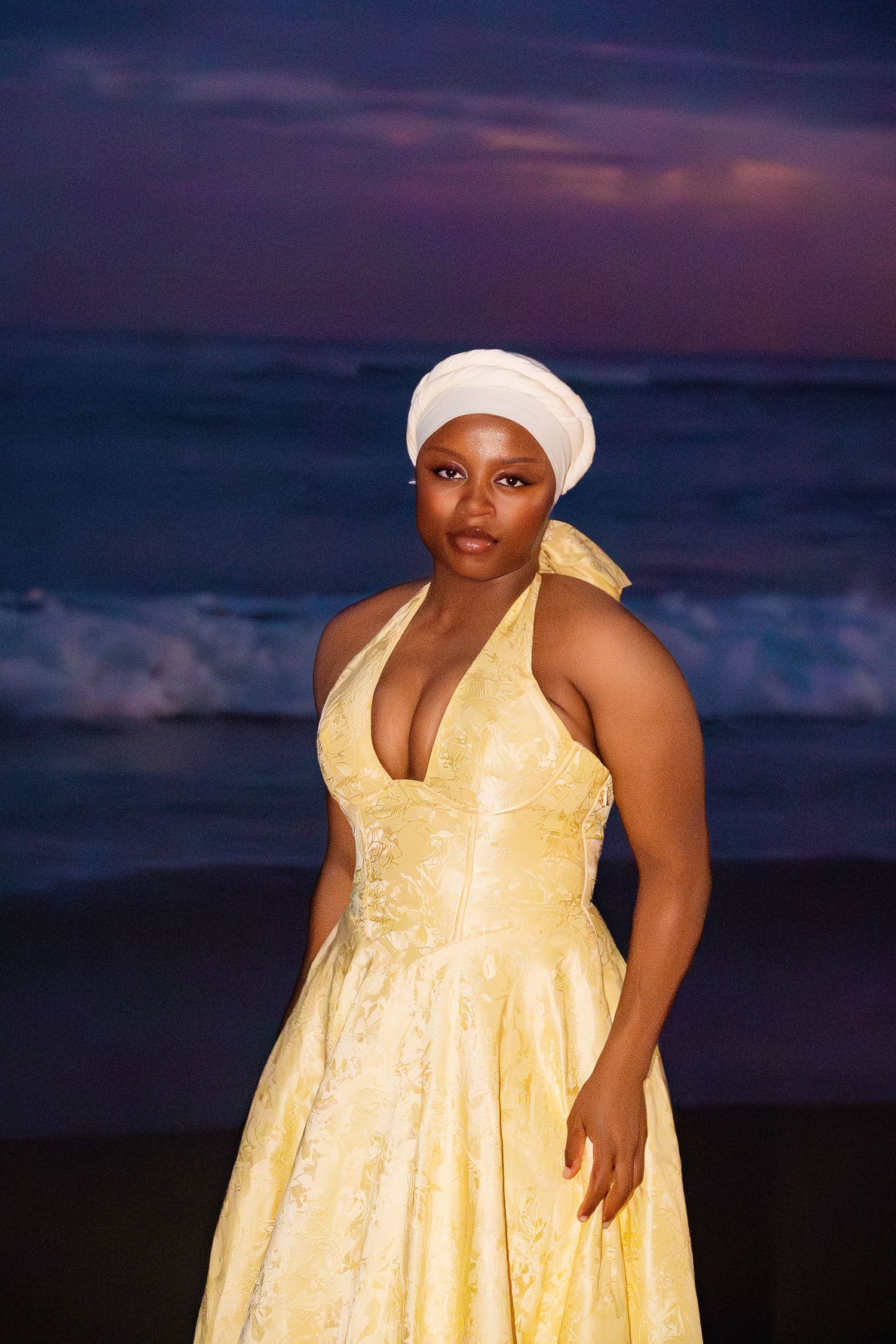 A woman in an elegant yellow halter gown and white turban stands gracefully on a beach at dusk.