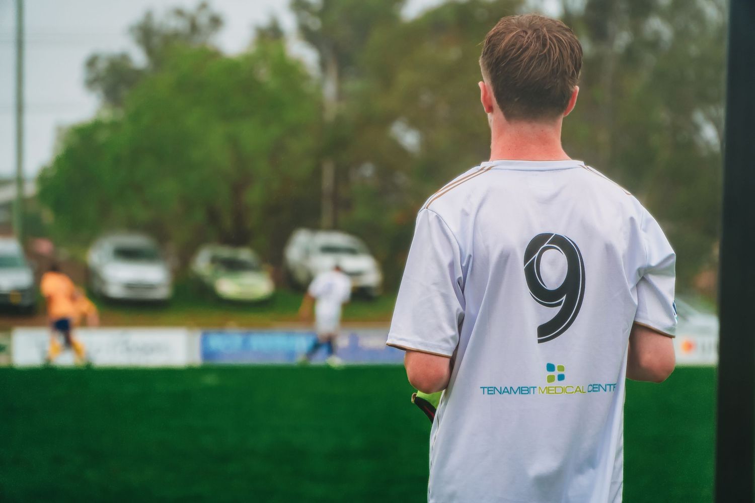 A soccer player wearing jersey number 9 stands on the sideline watching a match on a green field.