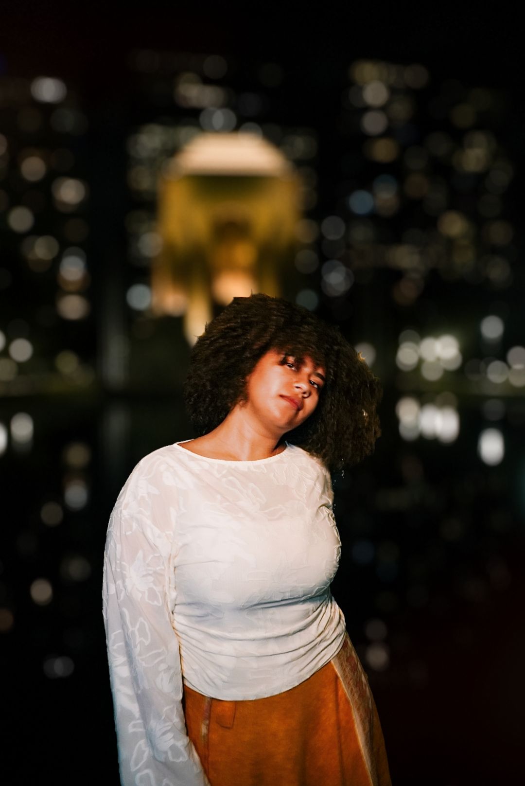 Woman with natural curly hair in white top and orange skirt posing with eyes closed at night outdoors.