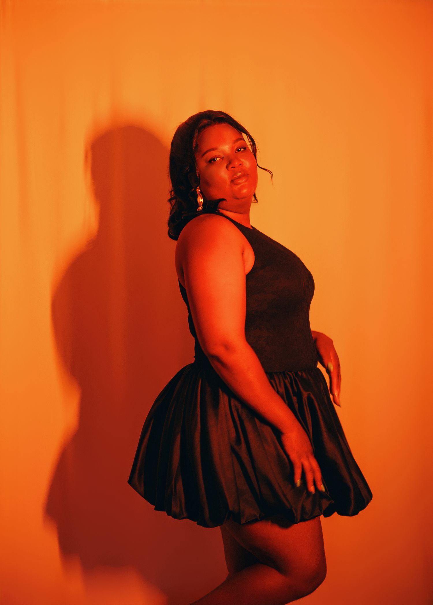 Woman in black bubble skirt dress posing sideways against orange background.