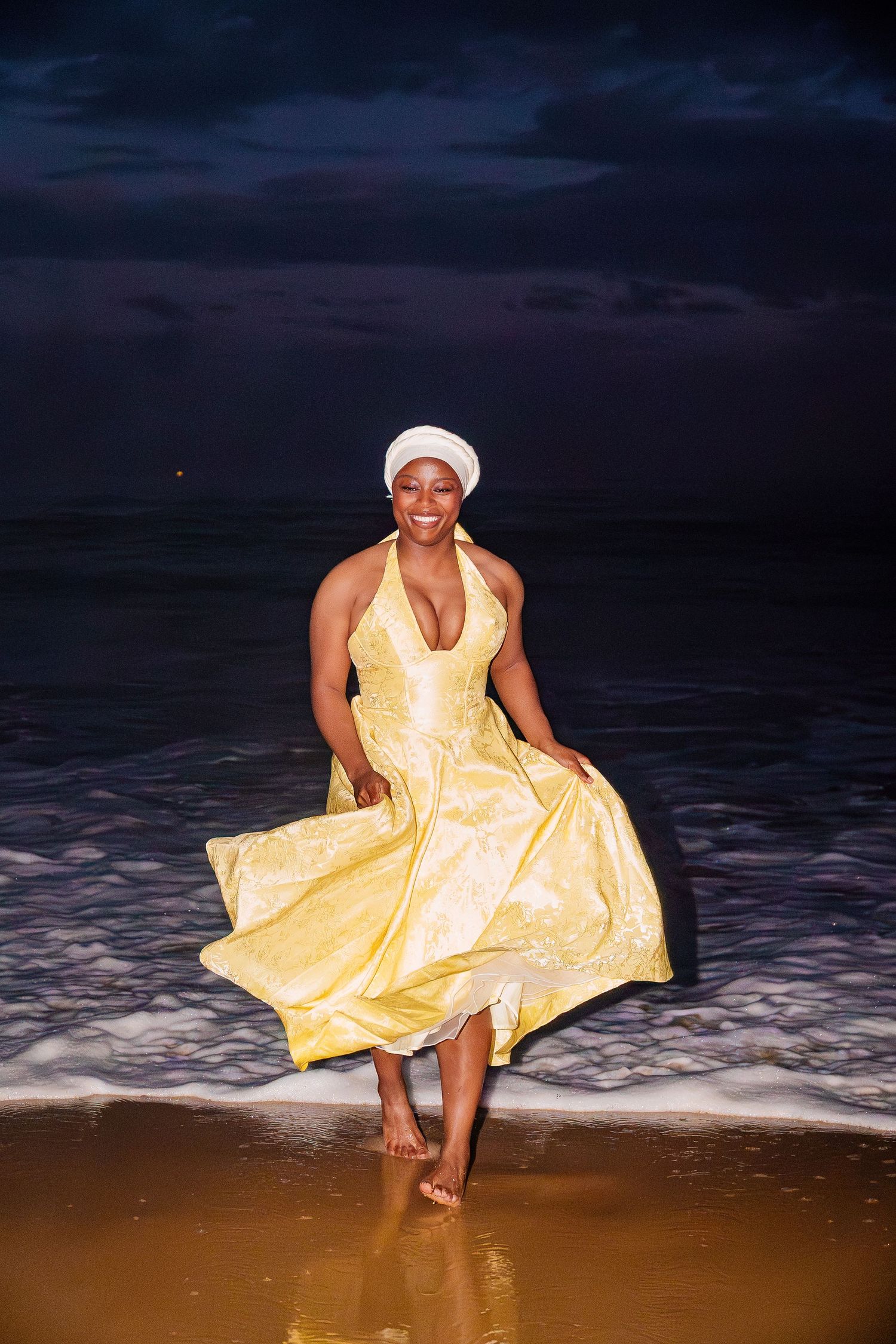 A woman in a flowing yellow halter dress and white turban walks barefoot through ocean waves at night.