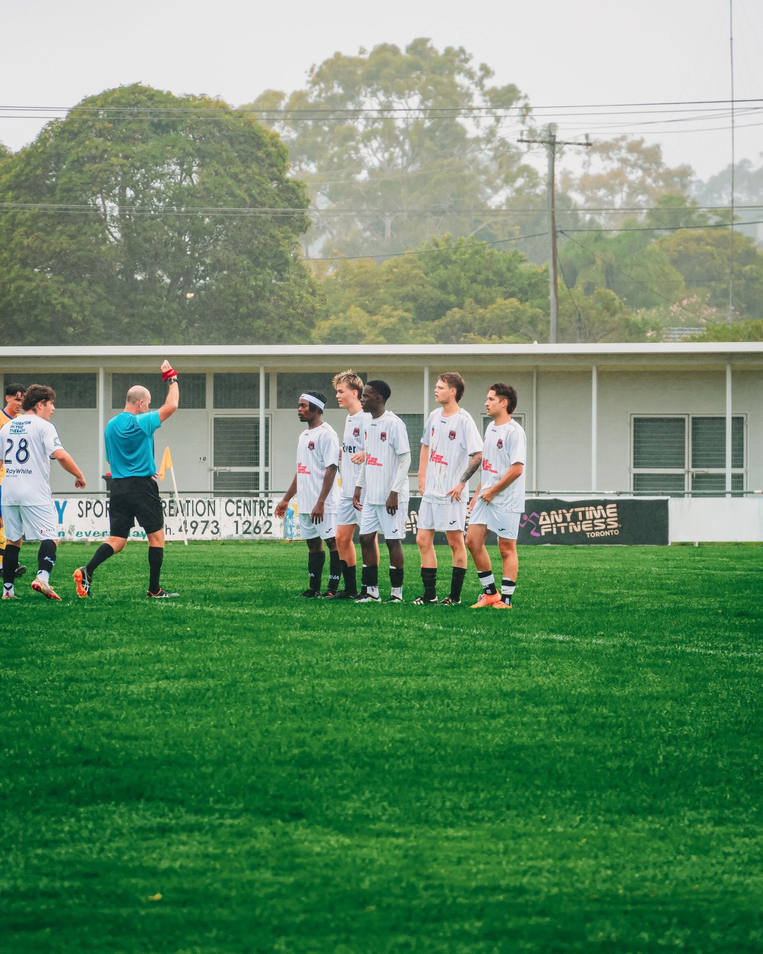 A referee shows a red card to soccer players on a green field with trees and buildings in the background.