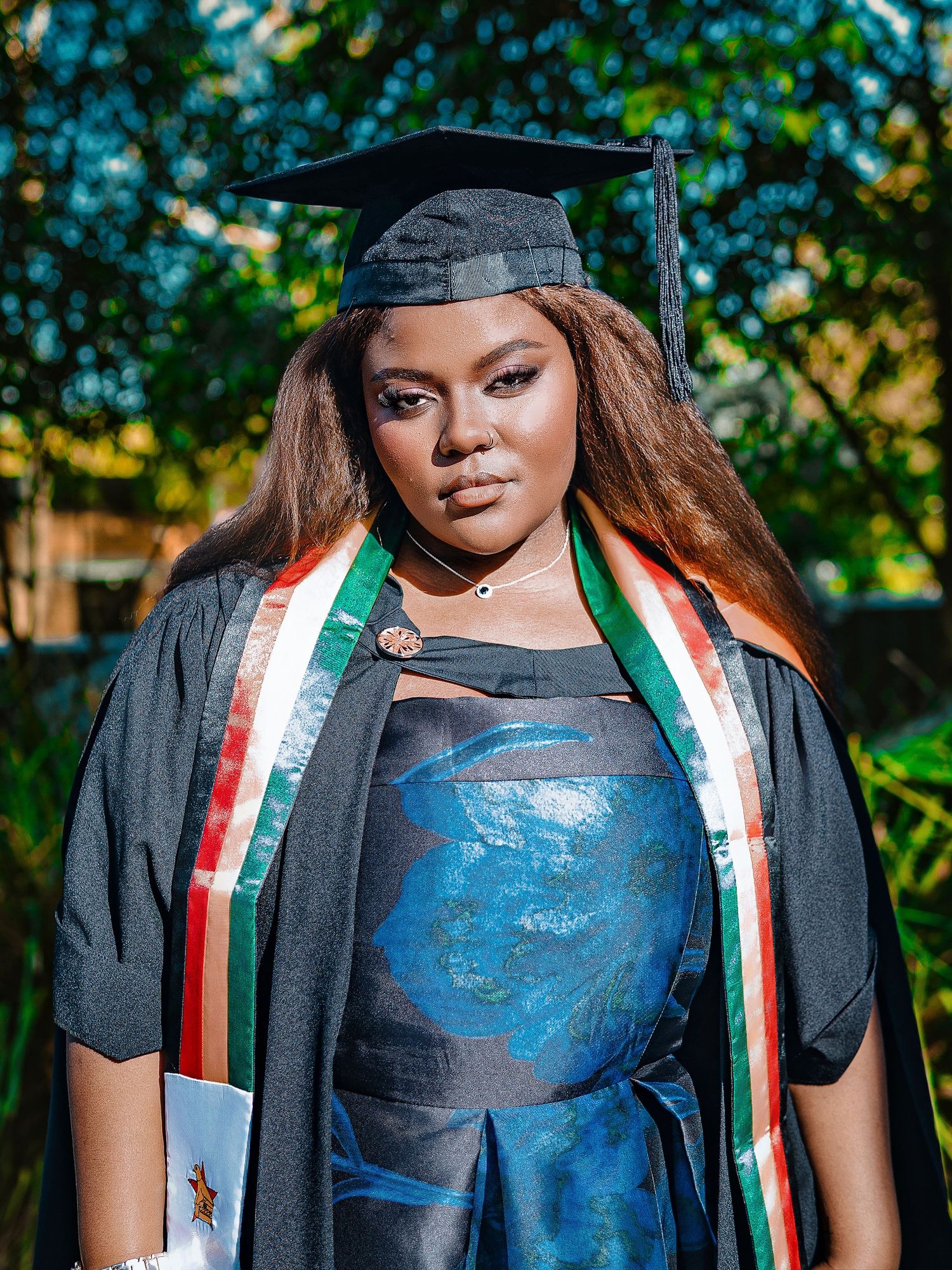 Graduate in cap, gown, and colorful stole poses outdoors.