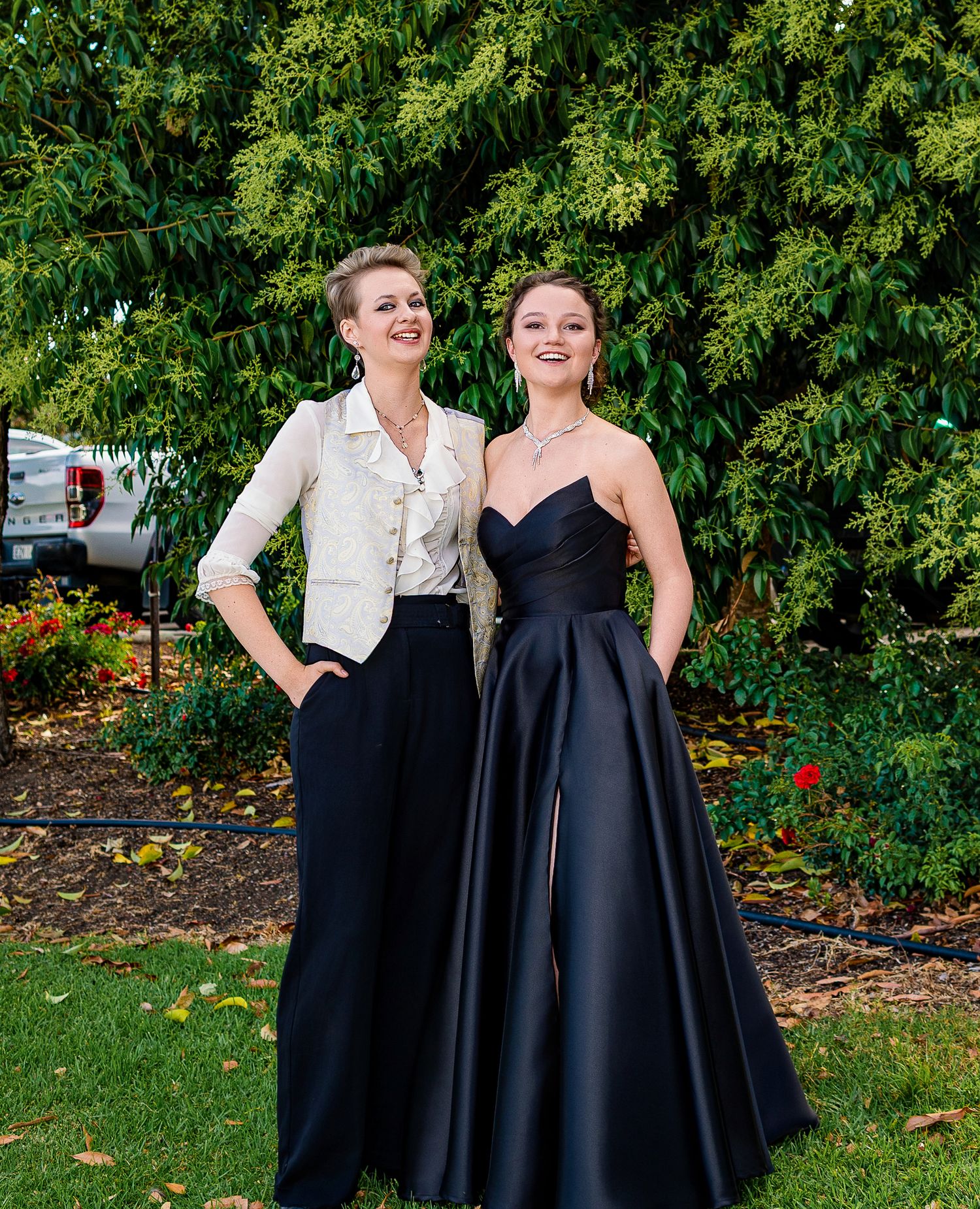 Two women in elegant formal wear pose outdoors, one in black gown and one in white jacket with wide-leg pants.