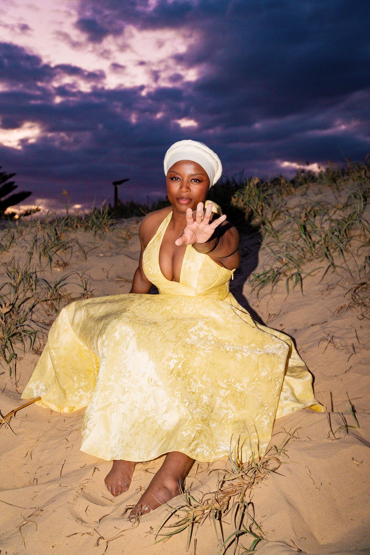 A woman in a yellow halter dress and white turban sits on sand dunes gesturing toward the camera at dusk.