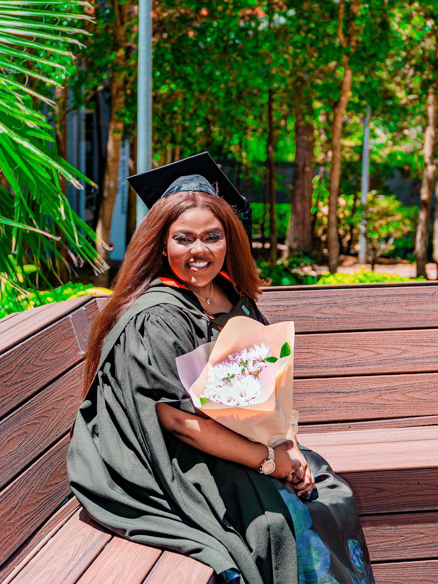 A smiling graduate in black cap and gown sits on a wooden bench holding a bouquet of flowers outdoors.