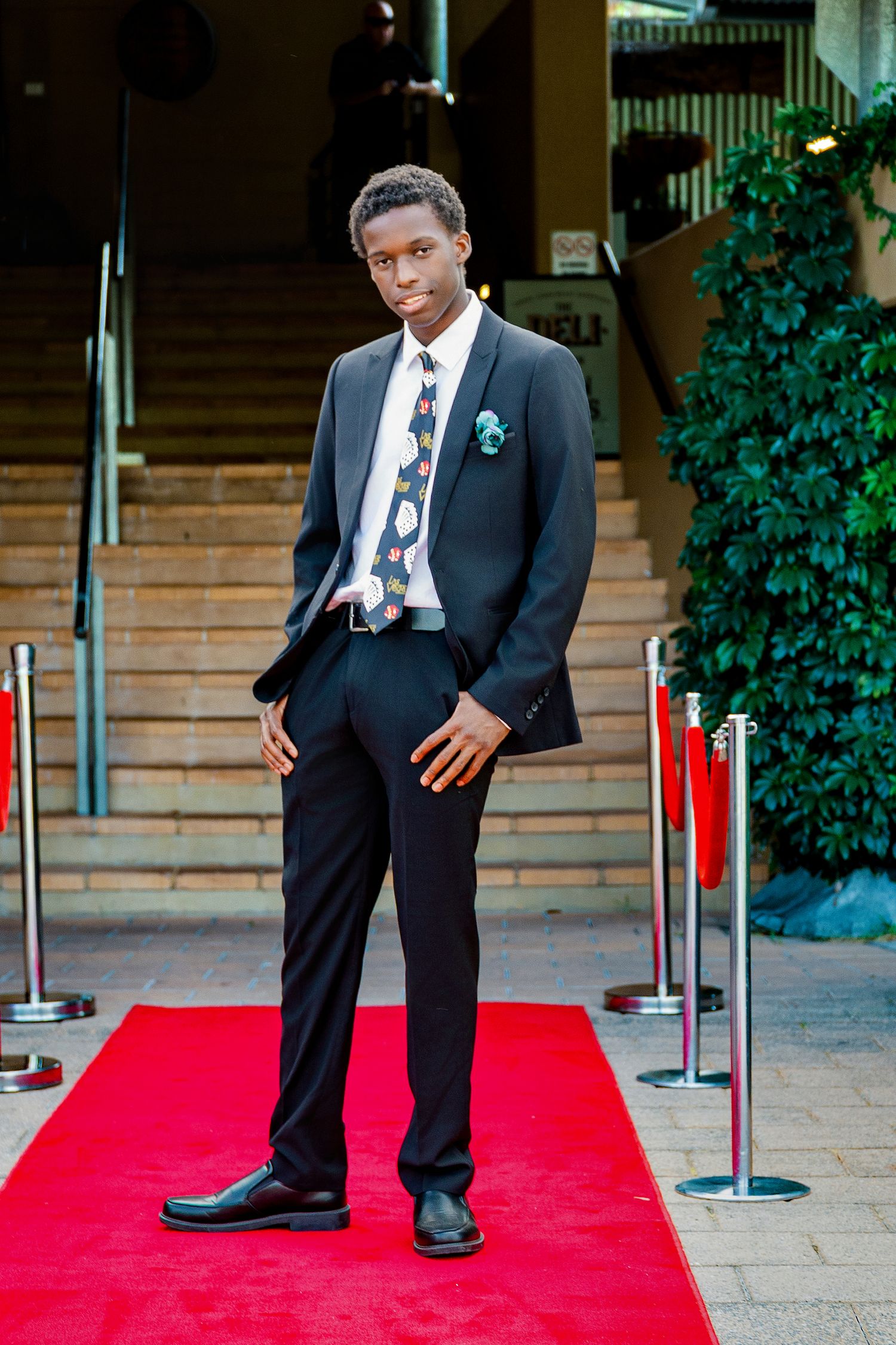 Young man in black suit with patterned tie posing confidently on red carpet.