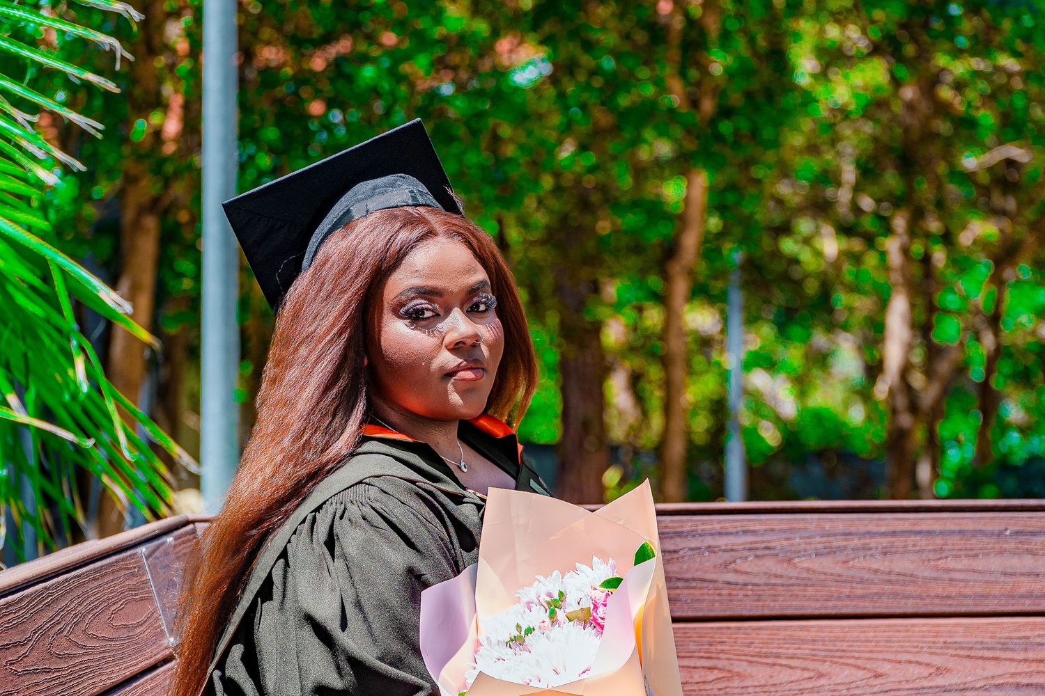 Young woman in graduation cap and gown holding a bouquet, smiling over her shoulder outdoors.