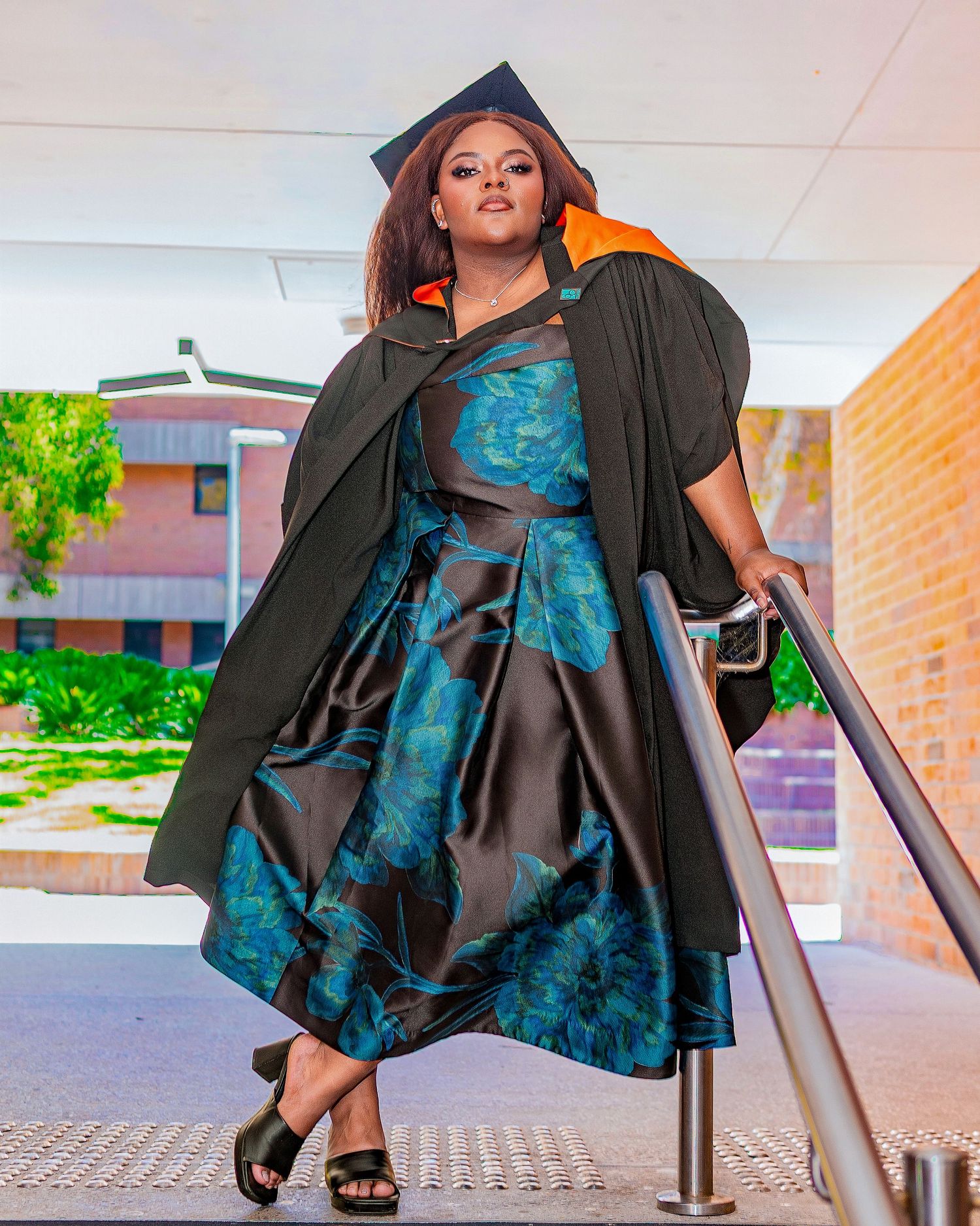 A graduate in black cap and gown with orange hood poses on stairs in a teal floral dress.