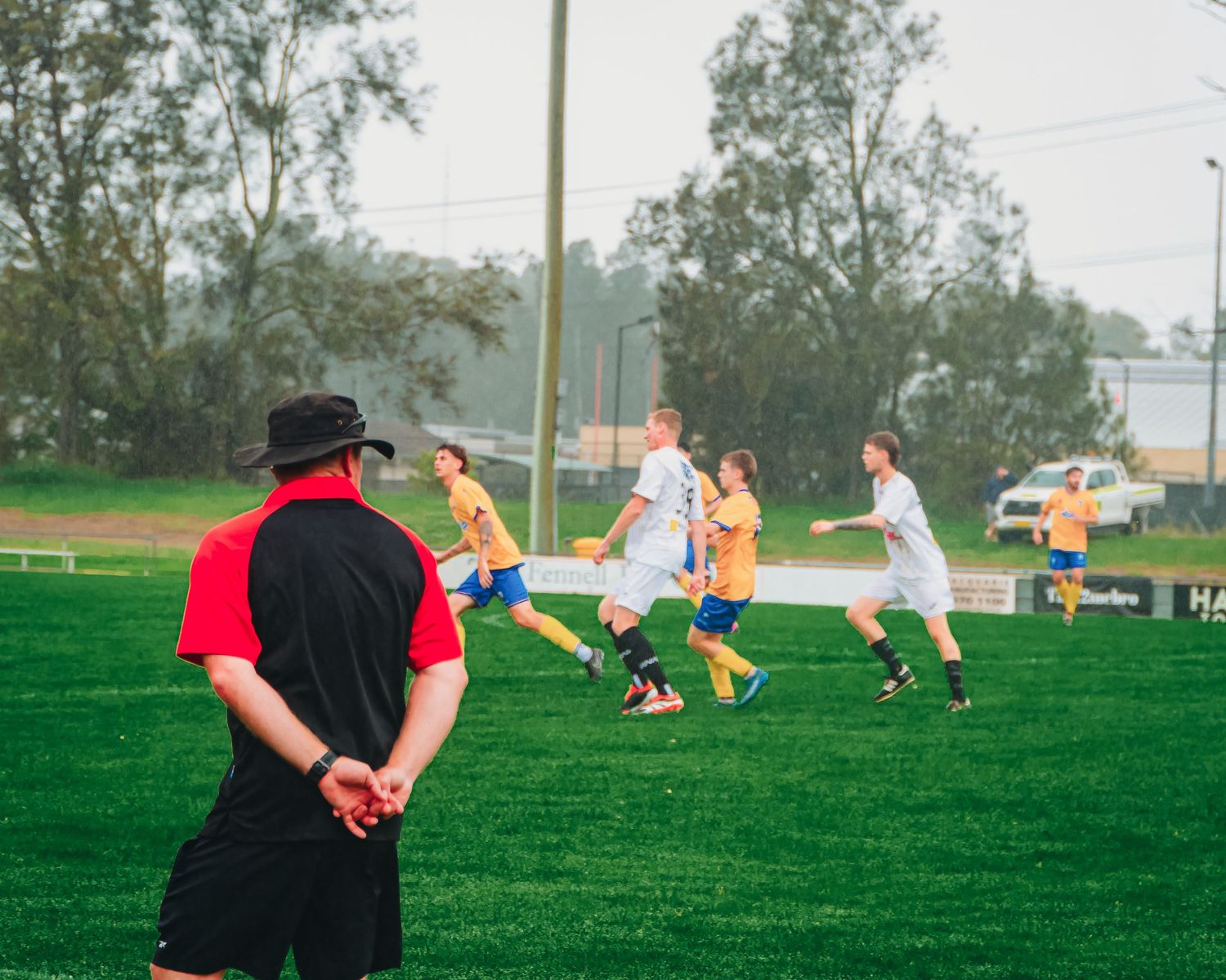 A referee in red and black watches youth soccer players in yellow and white uniforms compete on a green field.