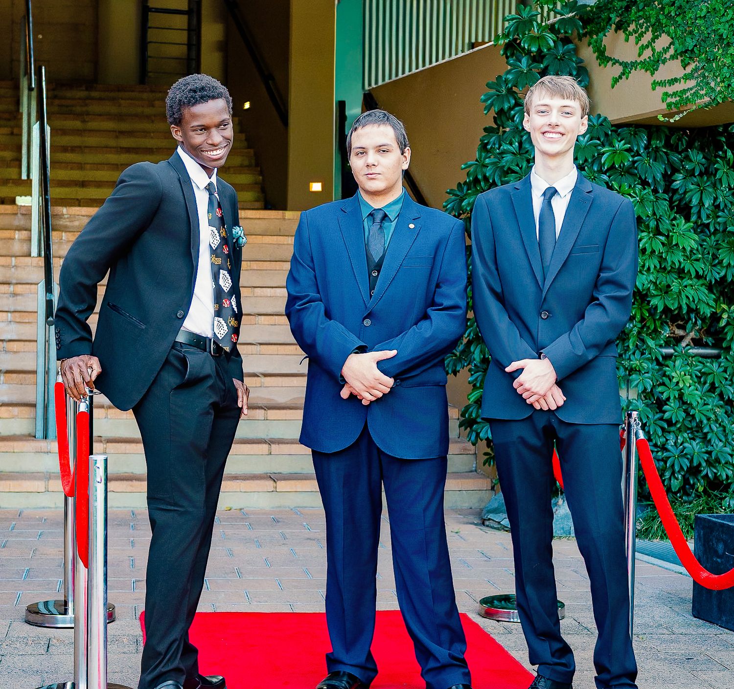 Three young men in formal suits pose on a red carpet at a formal event or gala.