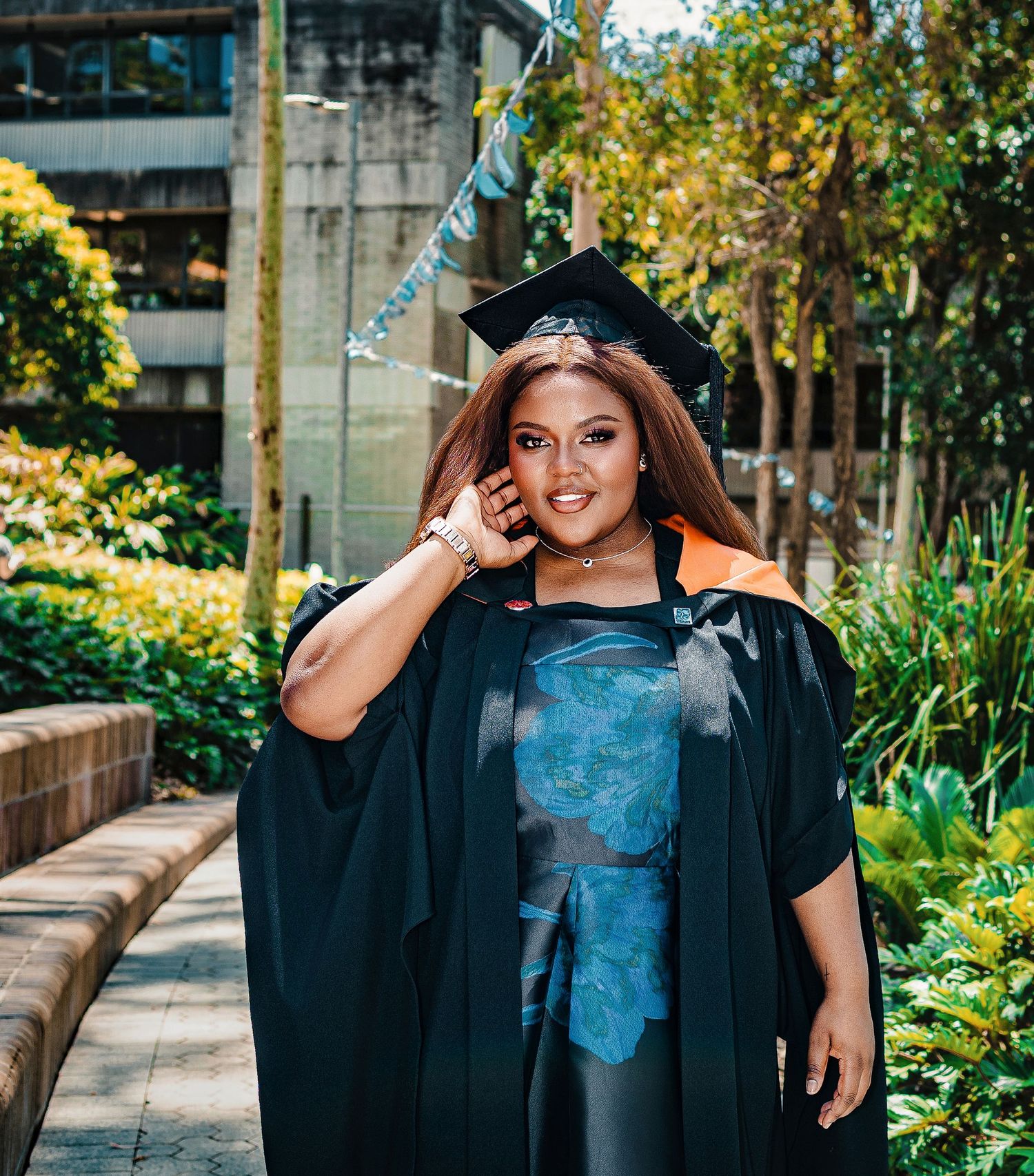 Woman in black graduation gown and cap smiling outdoors among lush greenery.