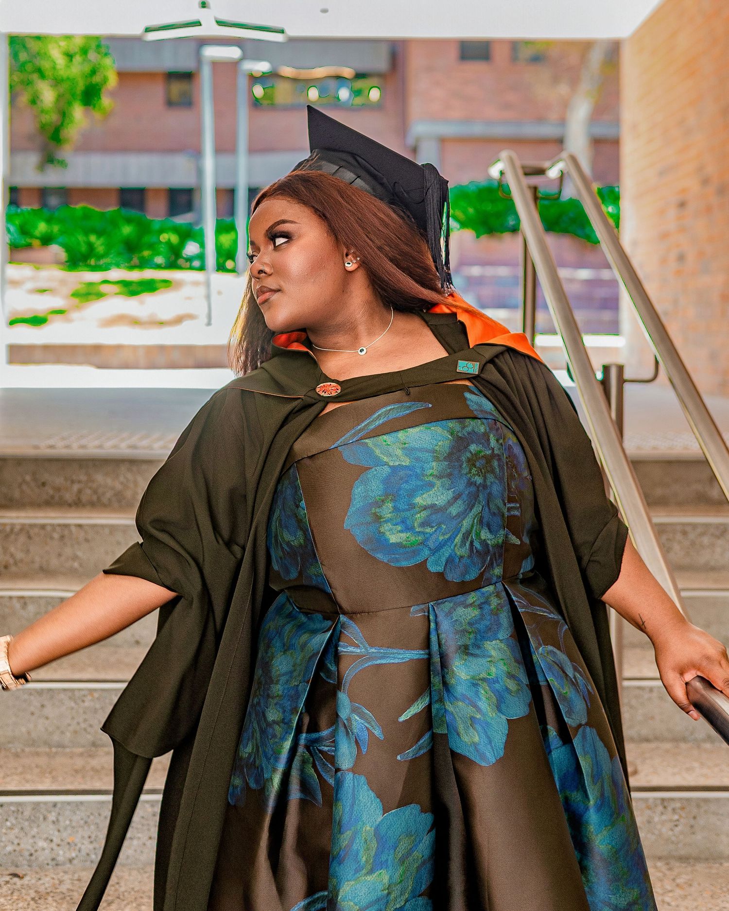 A woman in graduation cap, gown, and floral dress poses proudly on steps at her commencement ceremony.
