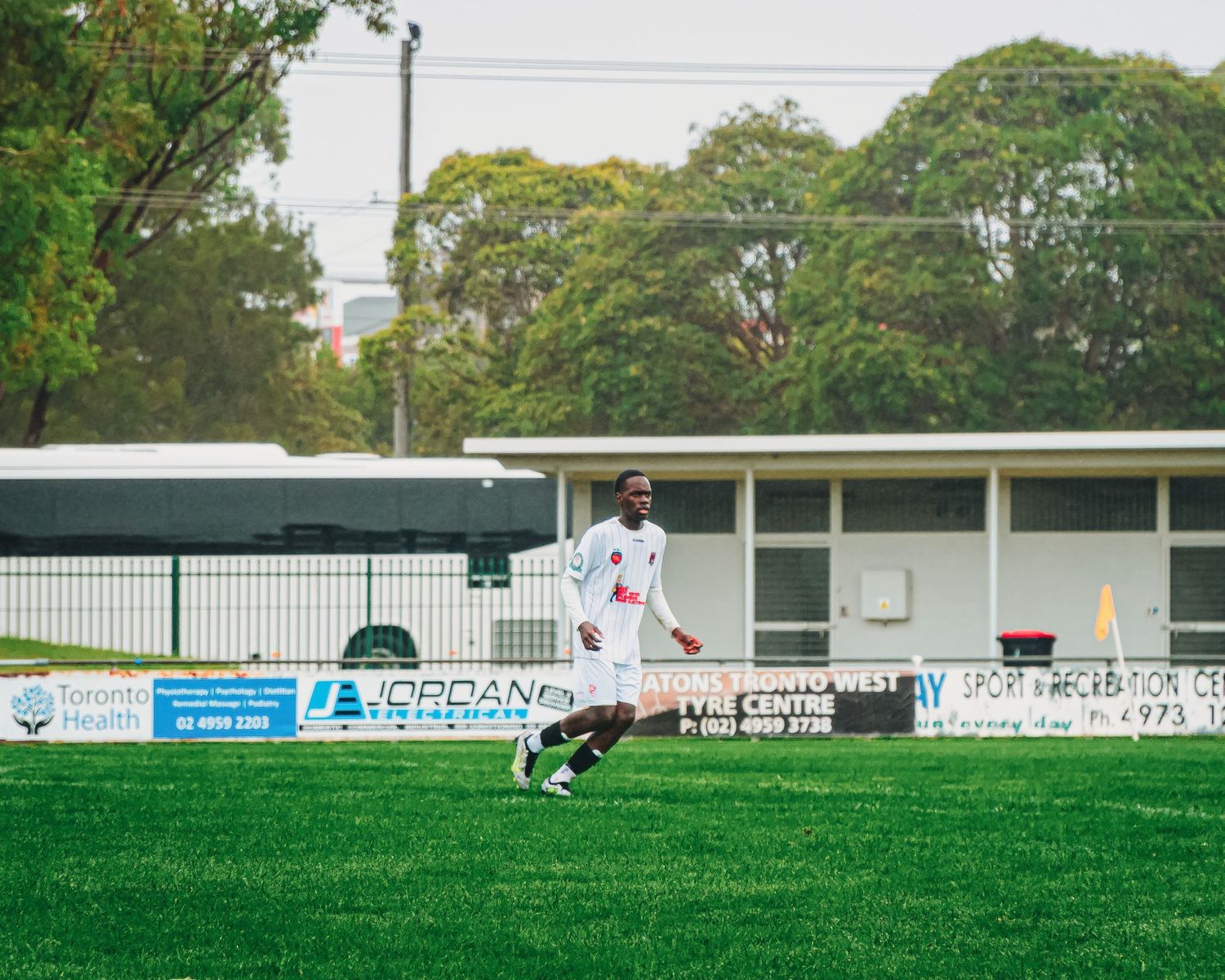 Soccer player in white uniform dribbling on green grass field with advertising boards and trees in background.