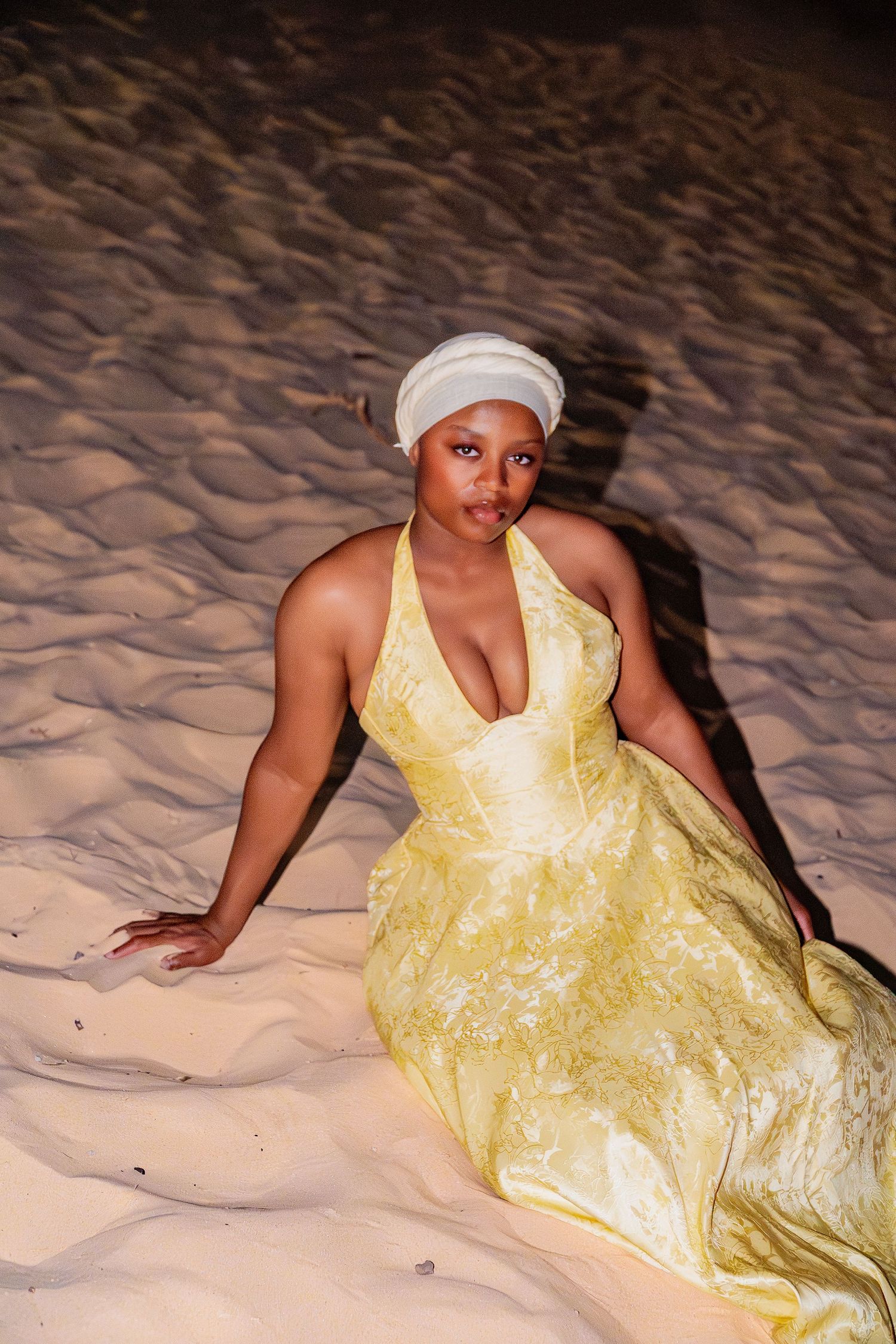 A woman in a yellow halter dress and white turban poses seated on illuminated sand dunes at night.