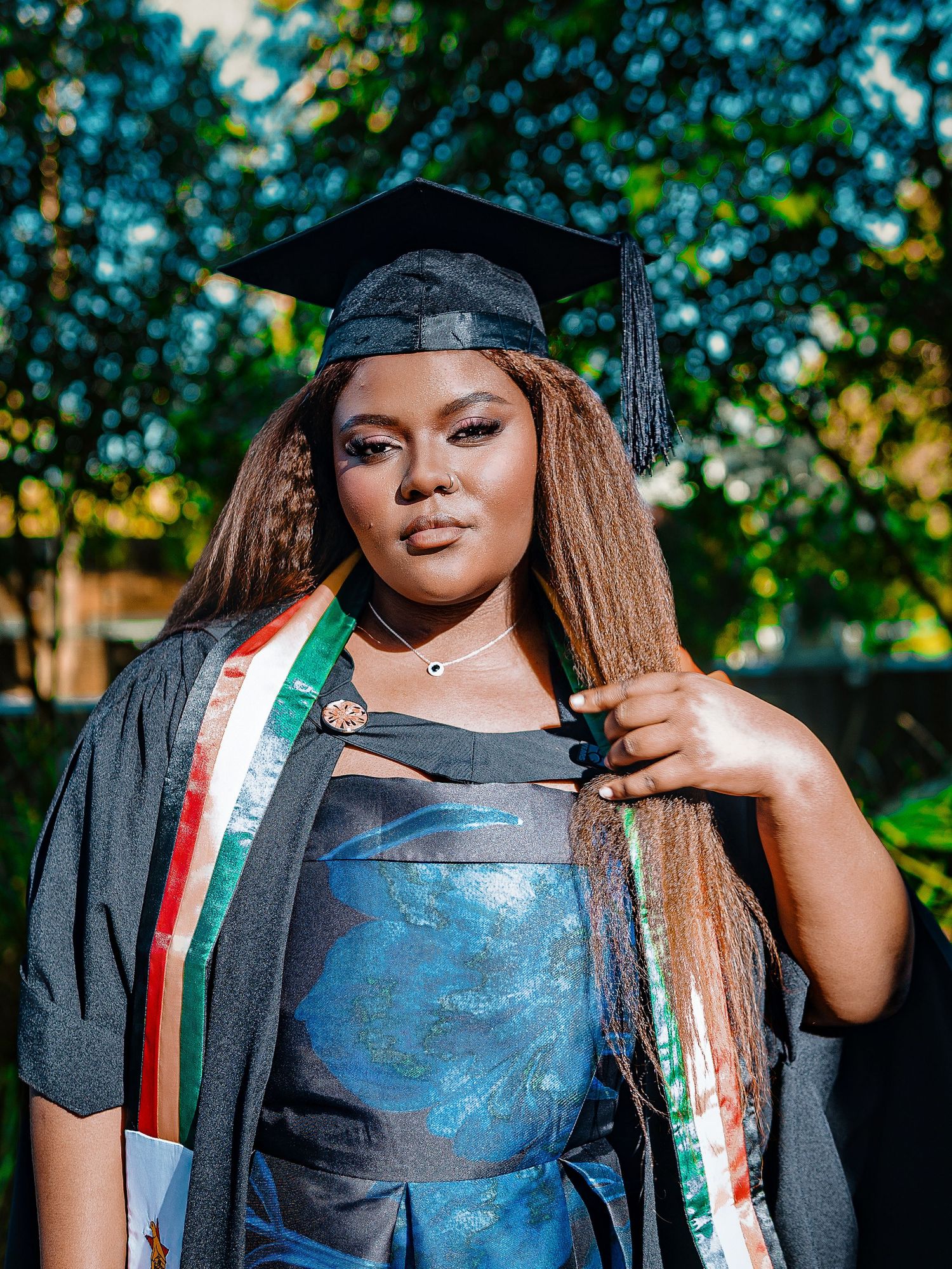 Graduate in black cap and gown with colorful sash holds braids while posing outdoors.