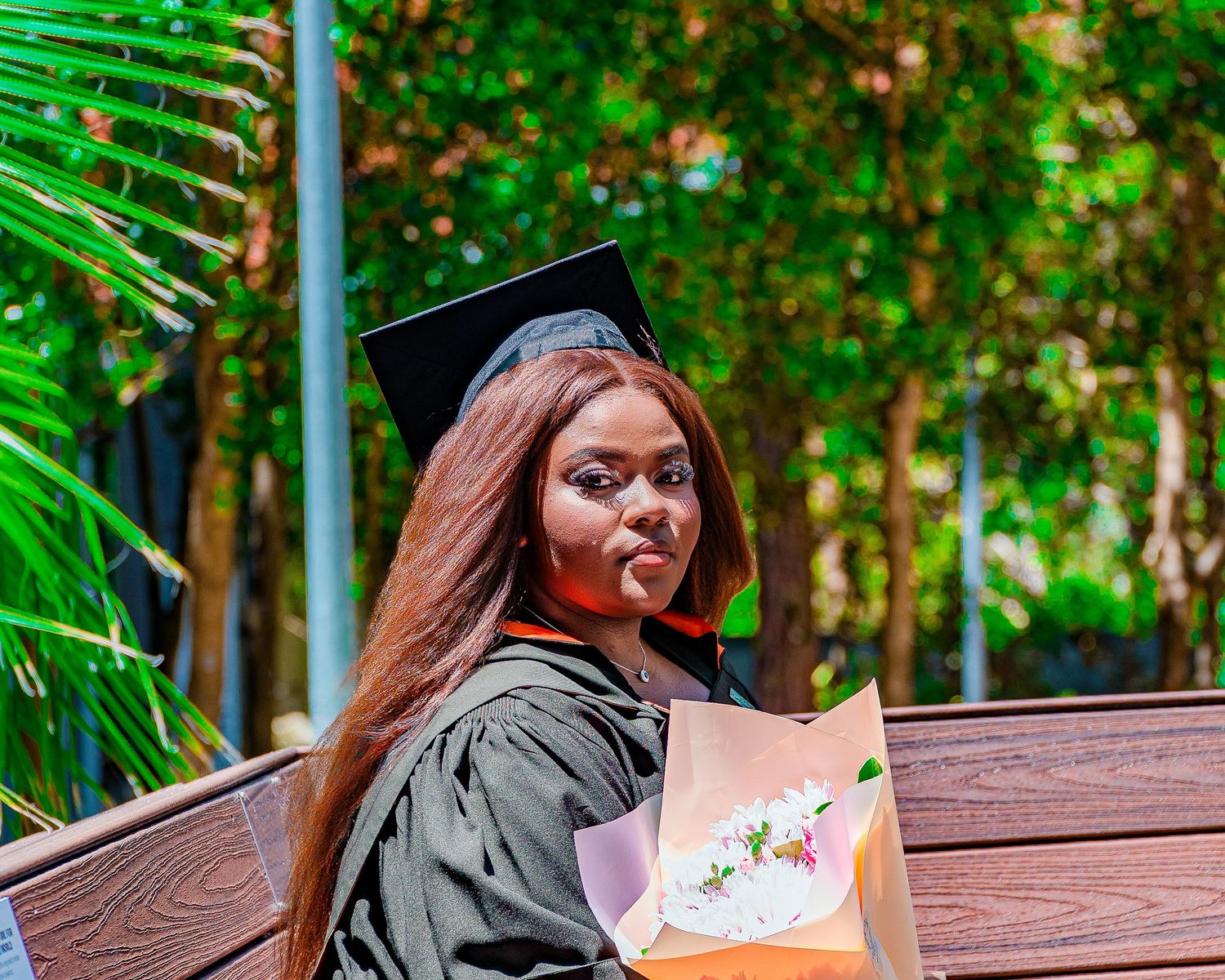 Young woman in graduation cap and gown smiling while holding a bouquet of flowers outdoors.