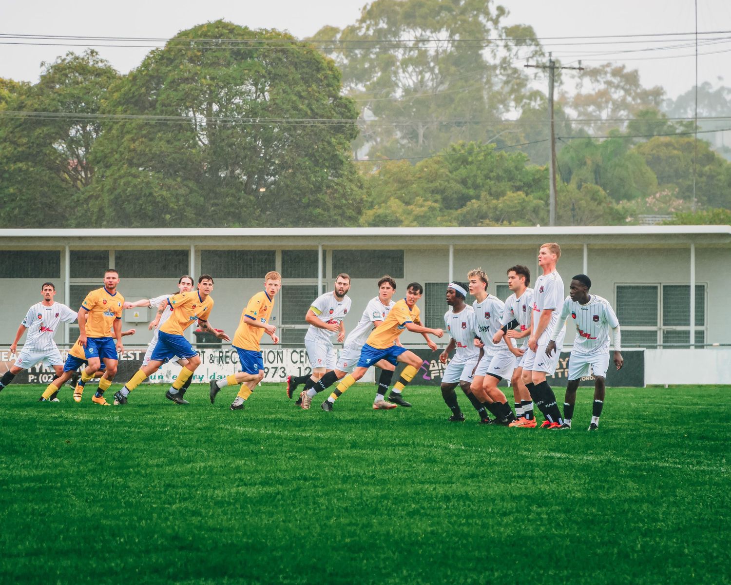Soccer players in yellow and white jerseys compete on a green field during a match with trees in the background.