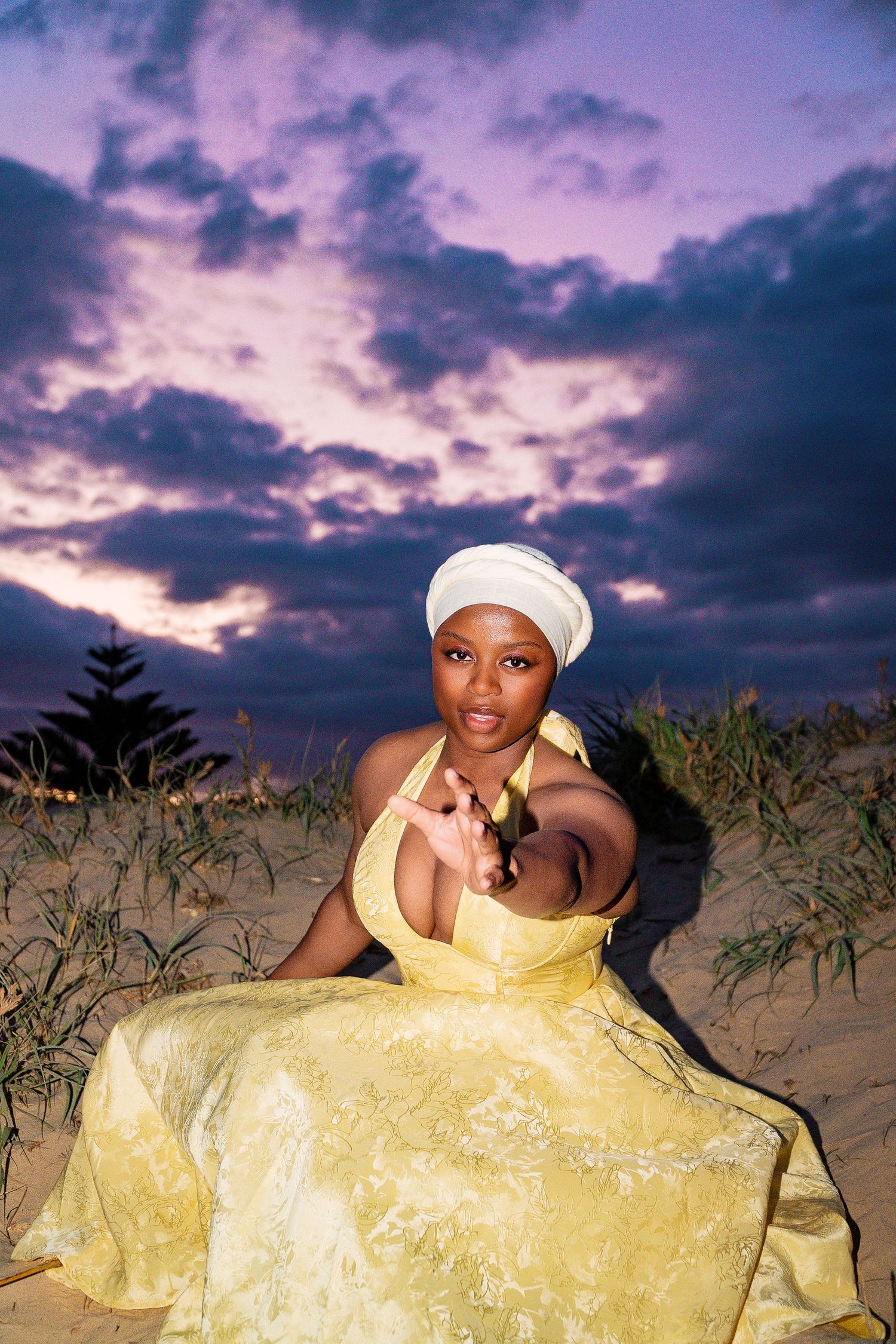 A woman in a yellow gown and white turban sits on sand dunes under a dramatic purple sunset sky.