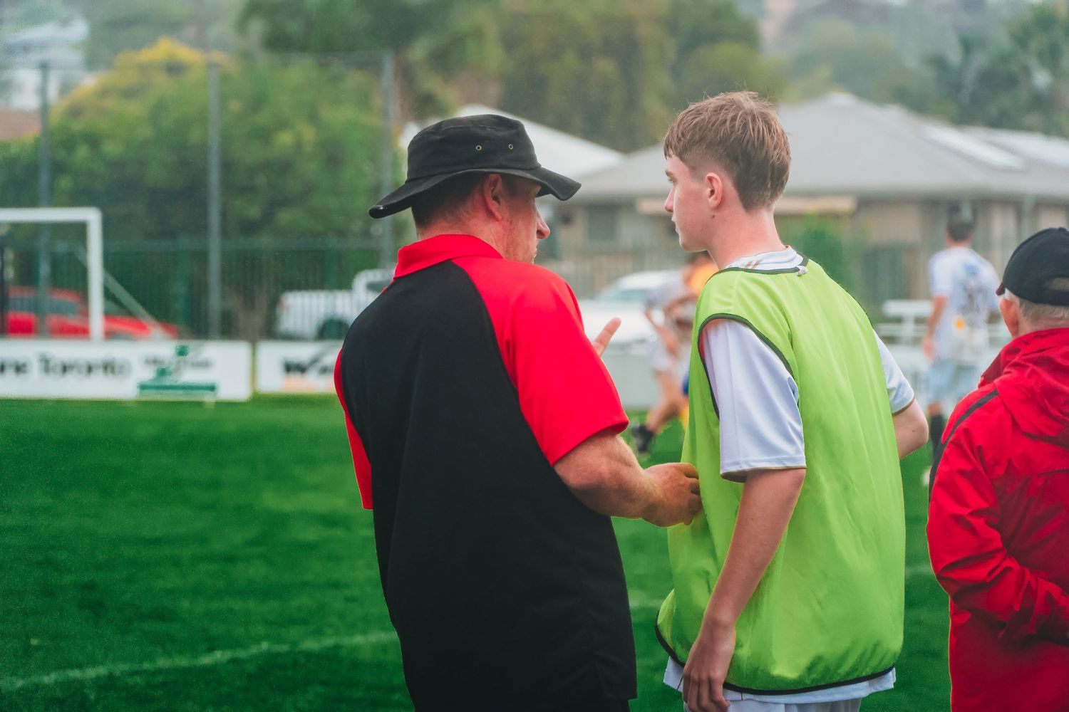 Soccer coach in black hat and red shirt giving instructions to young player in green vest on field.