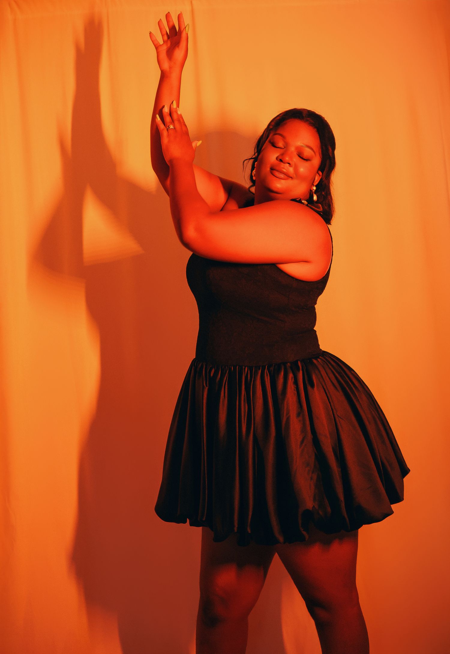 Woman in black strapless mini dress posing gracefully against orange-lit backdrop with arms raised.