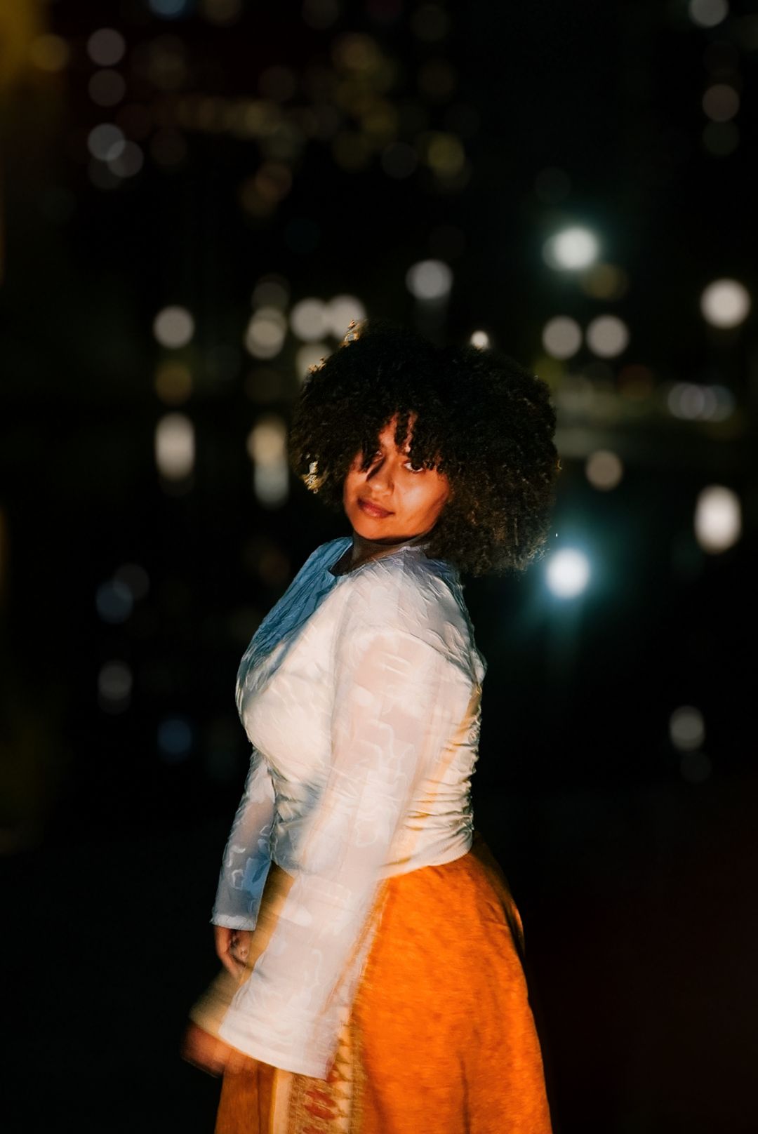 Woman in white lace top and orange skirt turning to look over her shoulder during a night photoshoot.