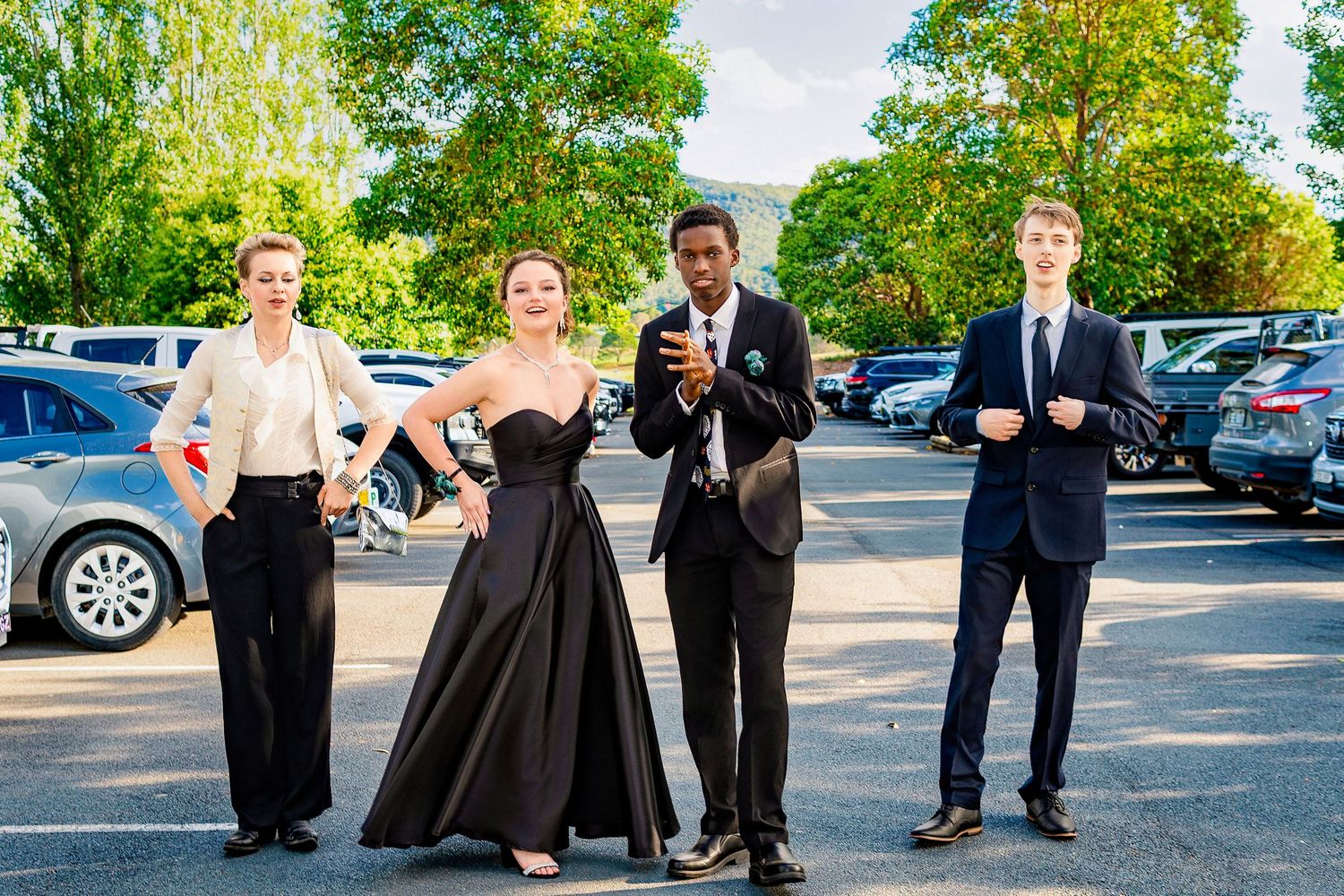 Four elegantly dressed teens pose in a parking lot, wearing formal attire including a black gown and suits.