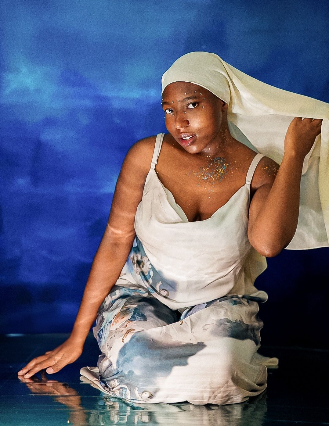 A woman in a white dress holds sheer fabric overhead, seated gracefully against a blue backdrop.