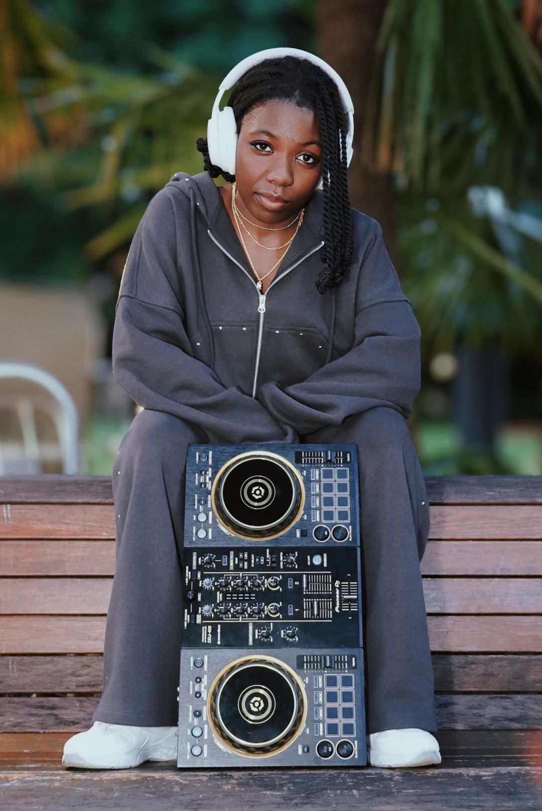 Young woman in gray hoodie sits on bench with DJ controller, wearing white headphones.